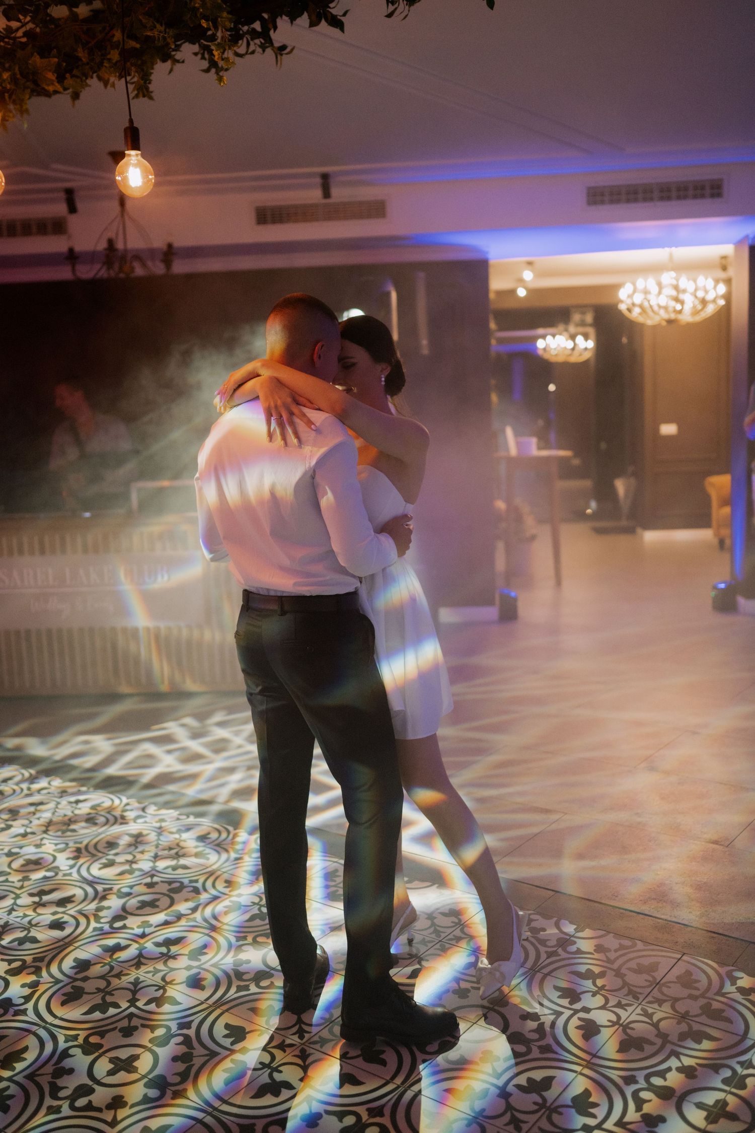 A couple shares a romantic dance on a patterned floor under moody lighting with hanging lights and chandeliers above.