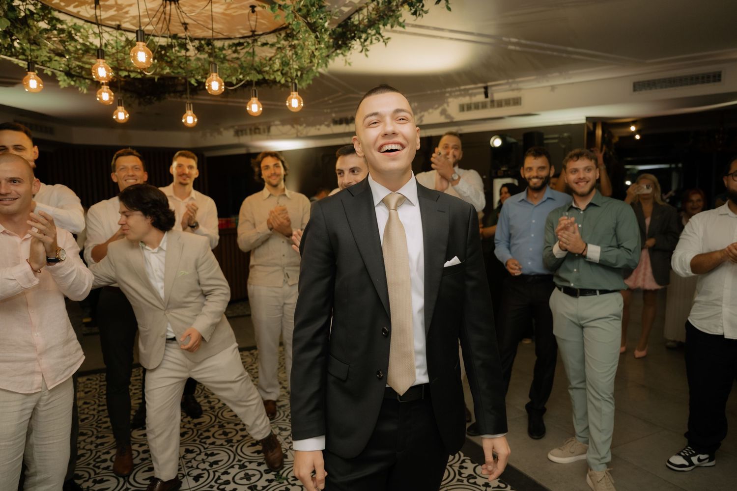 A group of wedding guests dance and celebrate on a dimly lit dance floor under hanging string lights.