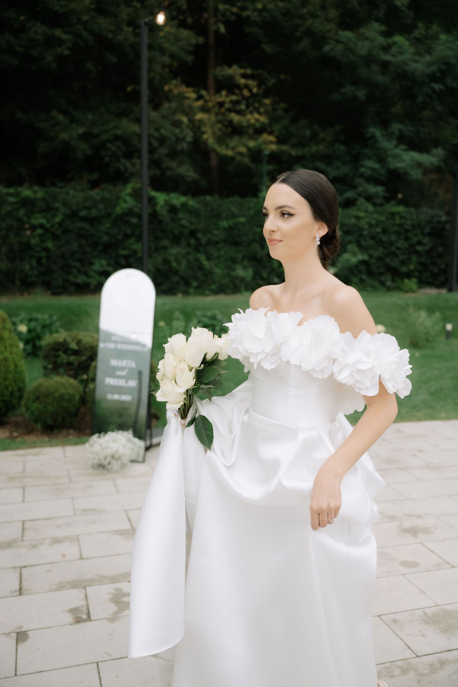A person in an elegant off-shoulder white wedding gown holds a bouquet of white flowers while standing on stone pavement.