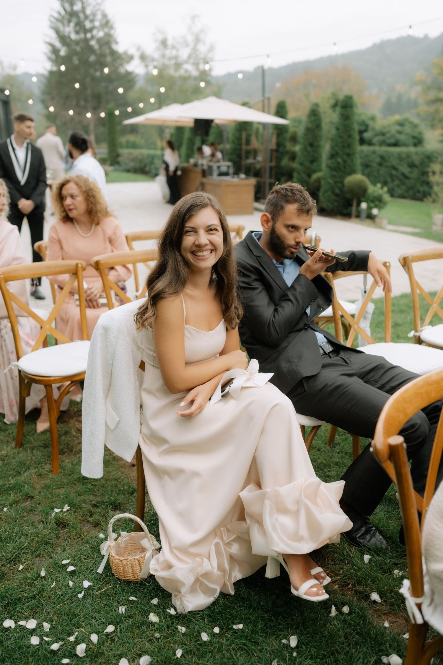 Wedding guests sitting on wooden chairs on a grassy lawn during an outdoor ceremony in the mountains.