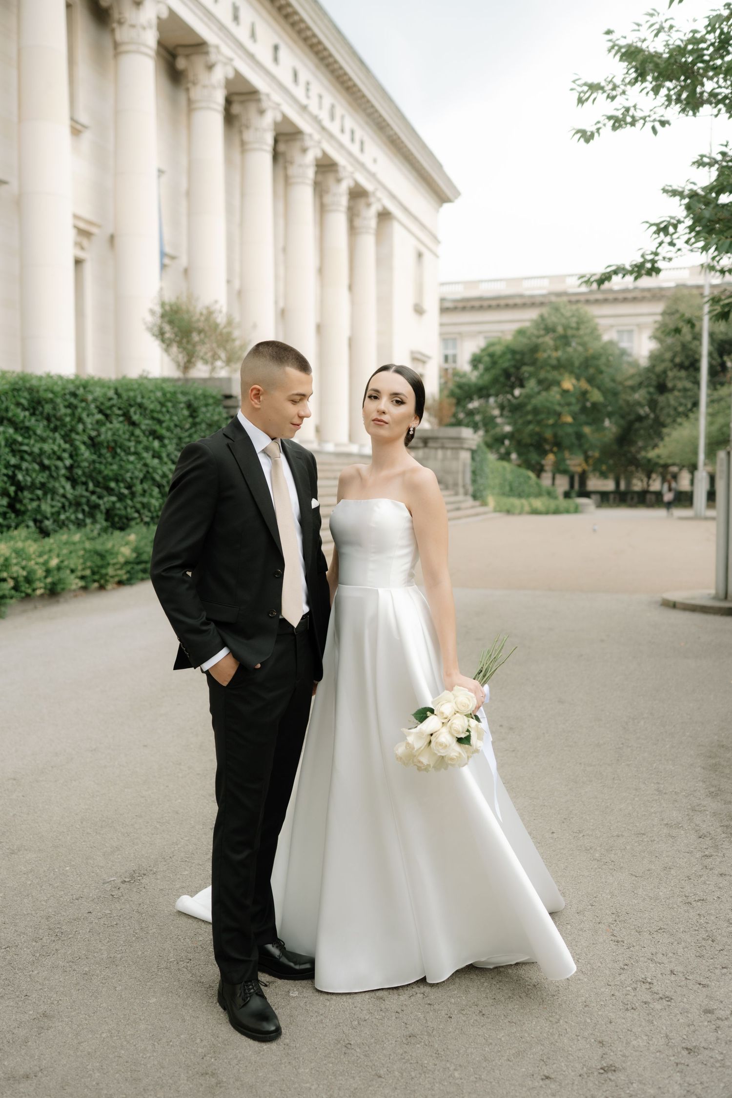 A wedding couple poses elegantly in front of a historic building with classical columns and architecture.