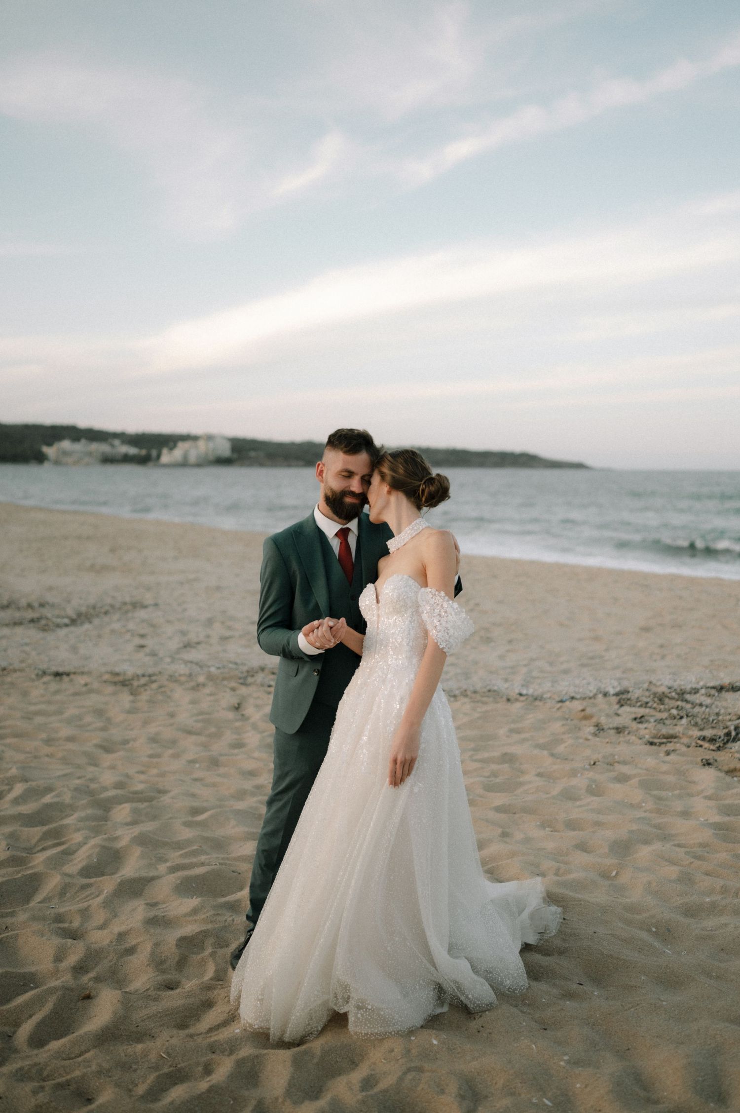 Bride and groom share a romantic moment on a sandy beach at dusk, with ocean waves and cliffs in the background.