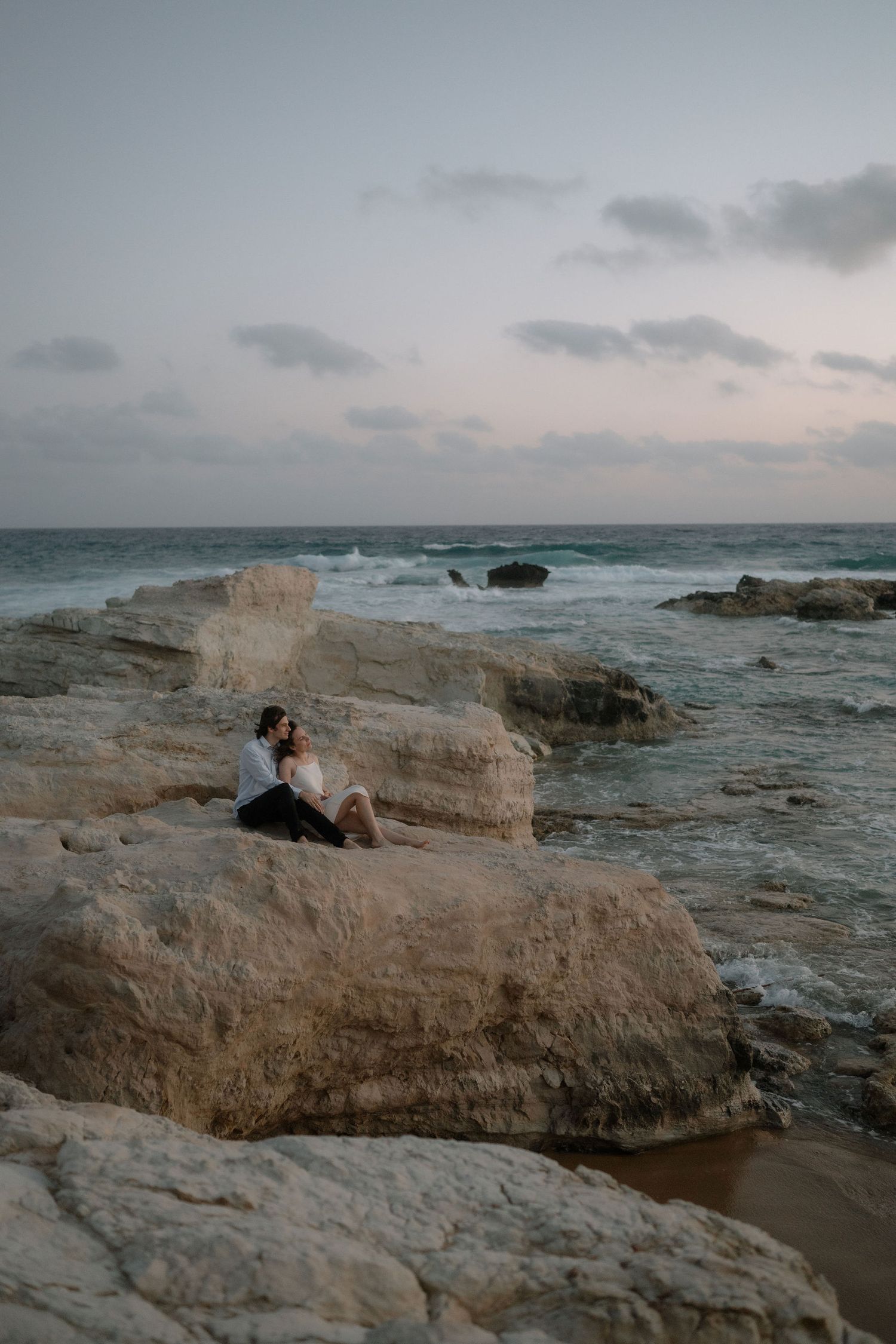 Couple sitting together on rocky coastal cliffs at sunset, with ocean waves and cloudy sky in the background.