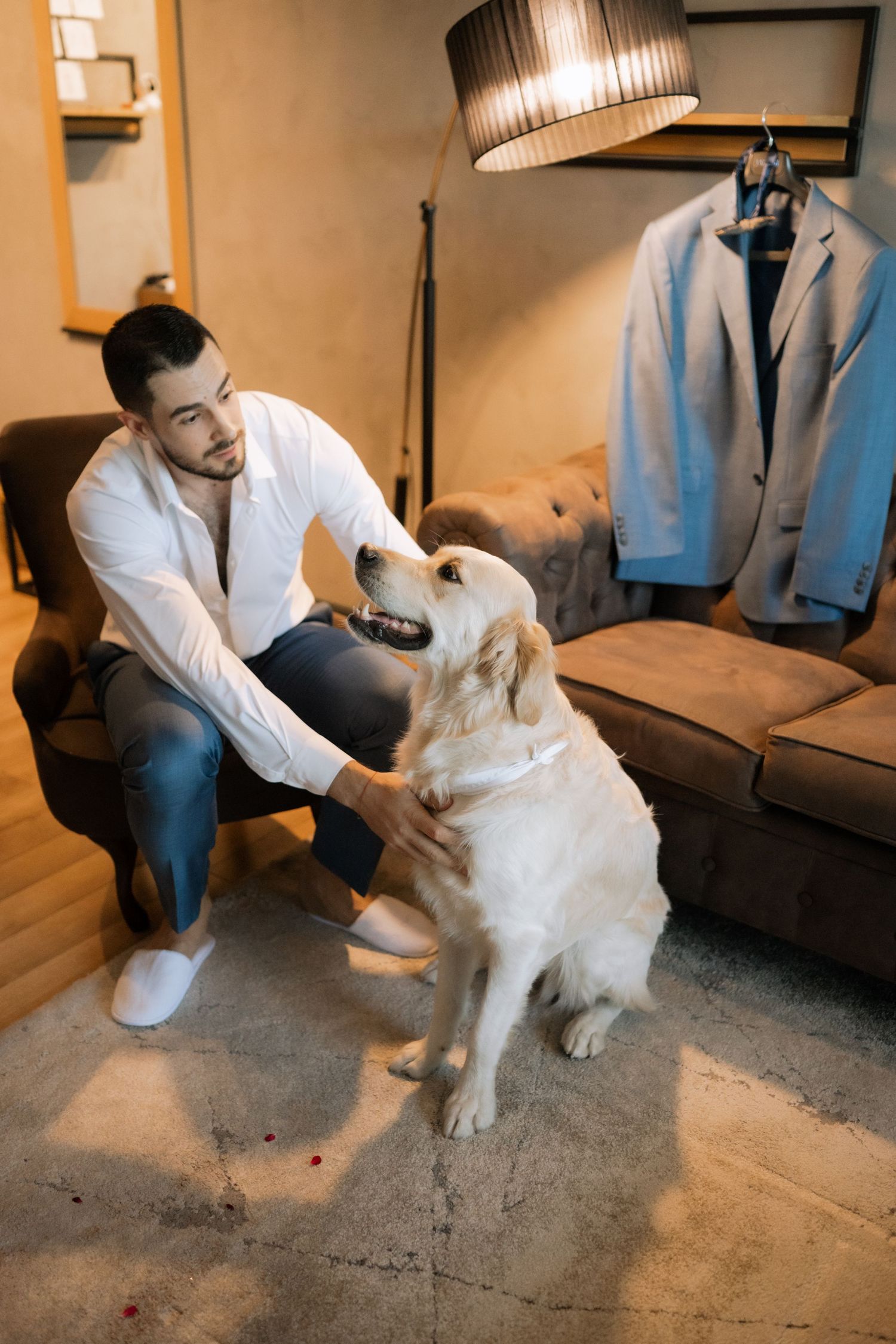 A person in a white shirt pets a friendly Golden Retriever while sitting on a couch with a blue jacket hanging nearby.