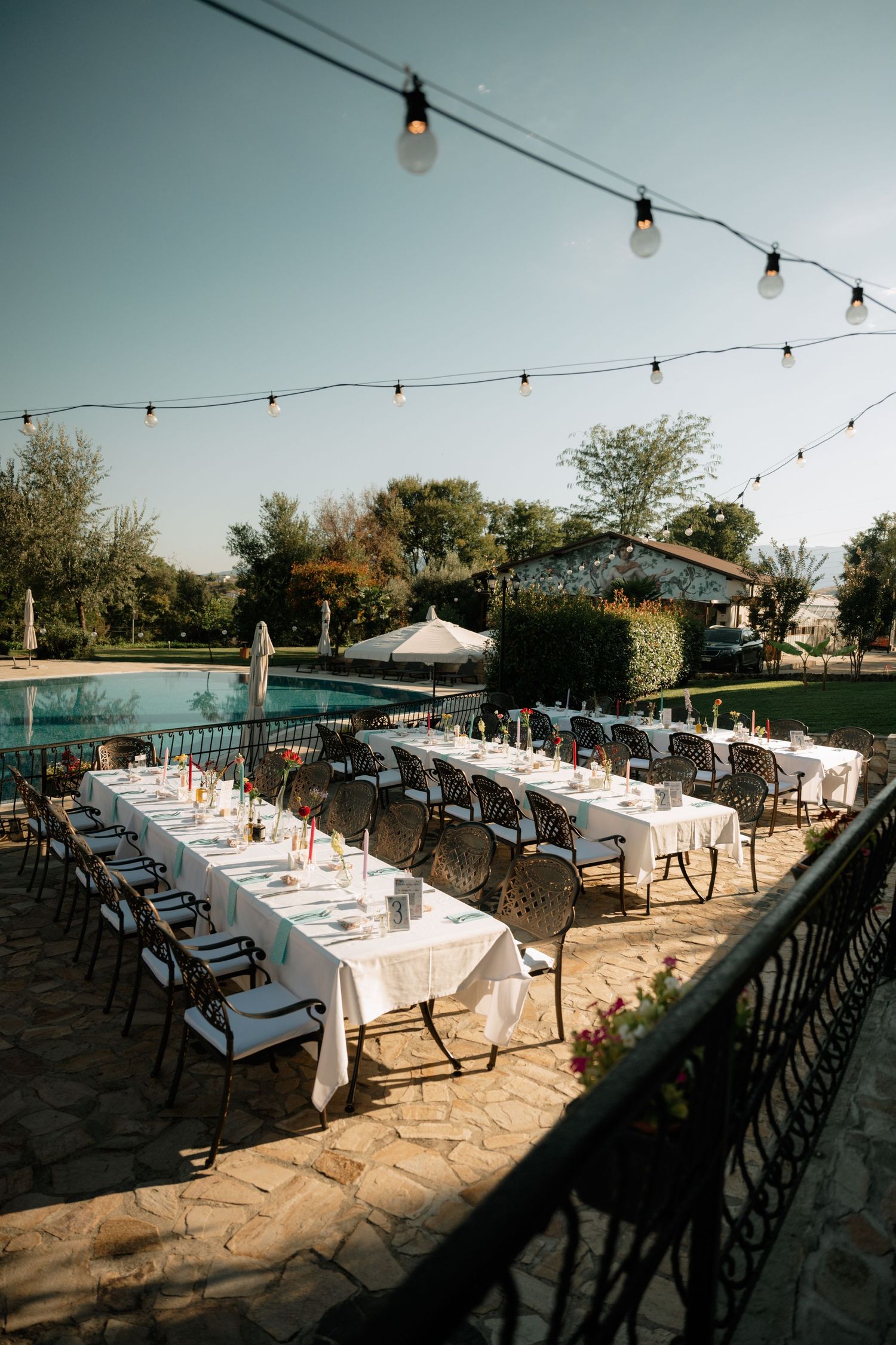 Outdoor dining setup with long tables under string lights on a stone patio at sunset creating an elegant atmosphere.