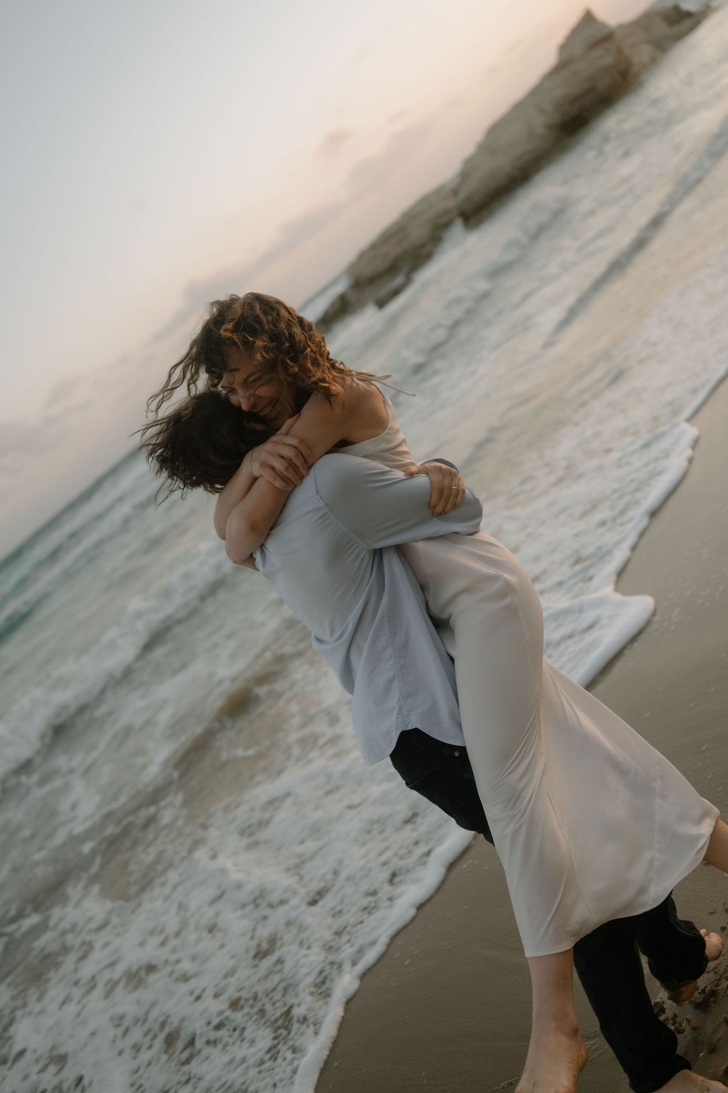 Couple embracing romantically on a beach at sunset, waves crashing behind them as man lifts woman in white dress.