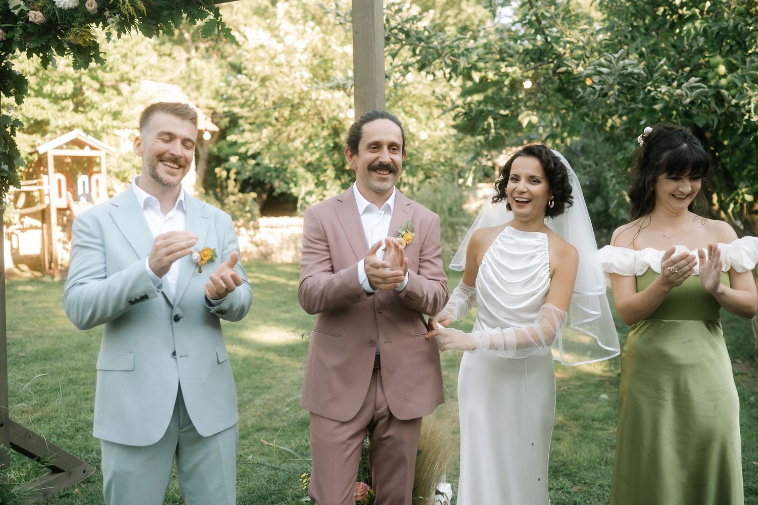 Wedding party members clap and dance together during an outdoor celebration in a garden setting.