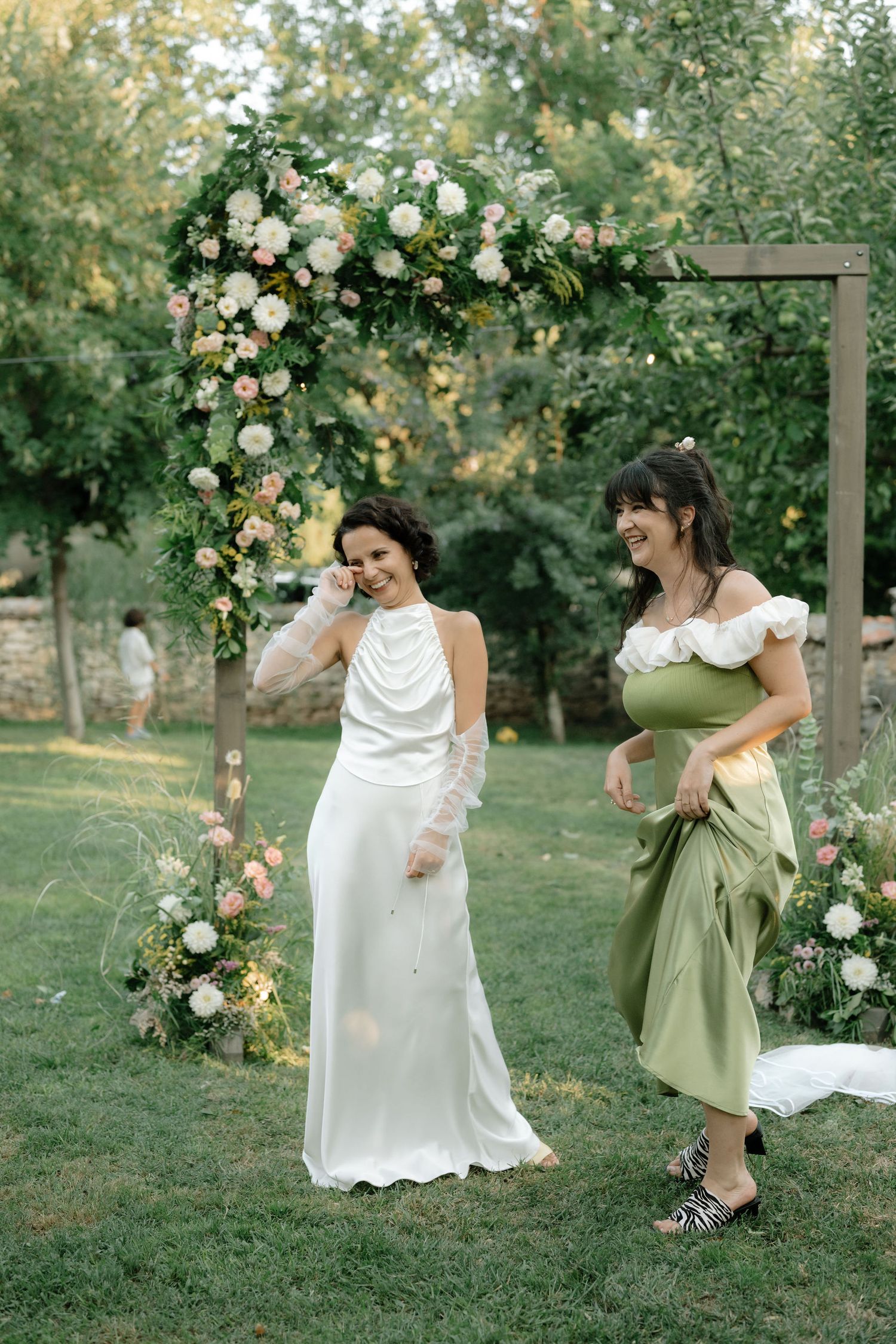 Two figures in white and green dresses dance playfully in a garden setting with blooming roses and trees.