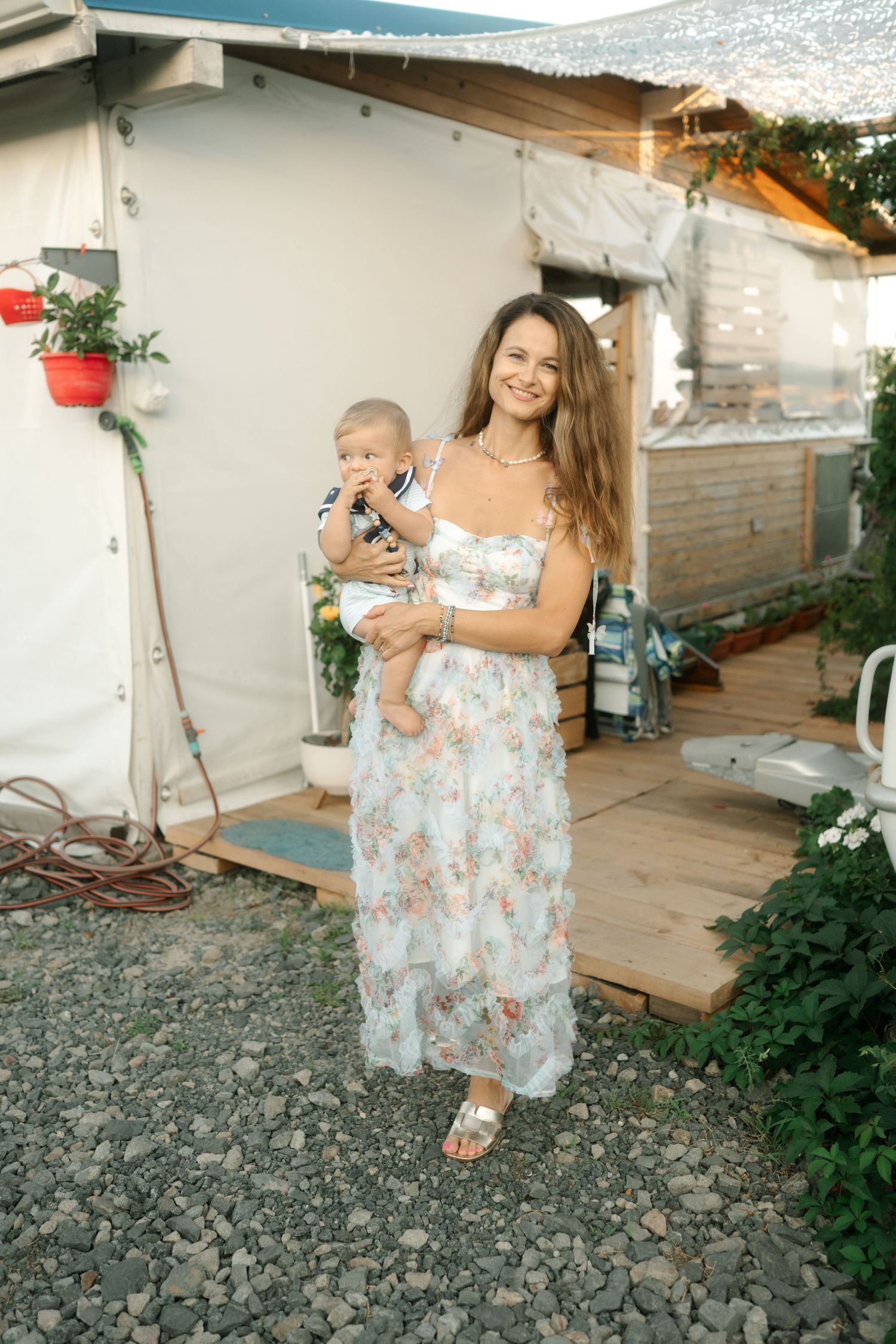 A mother in a floral sundress holds a baby while standing on gravel outside a rustic building in summer.