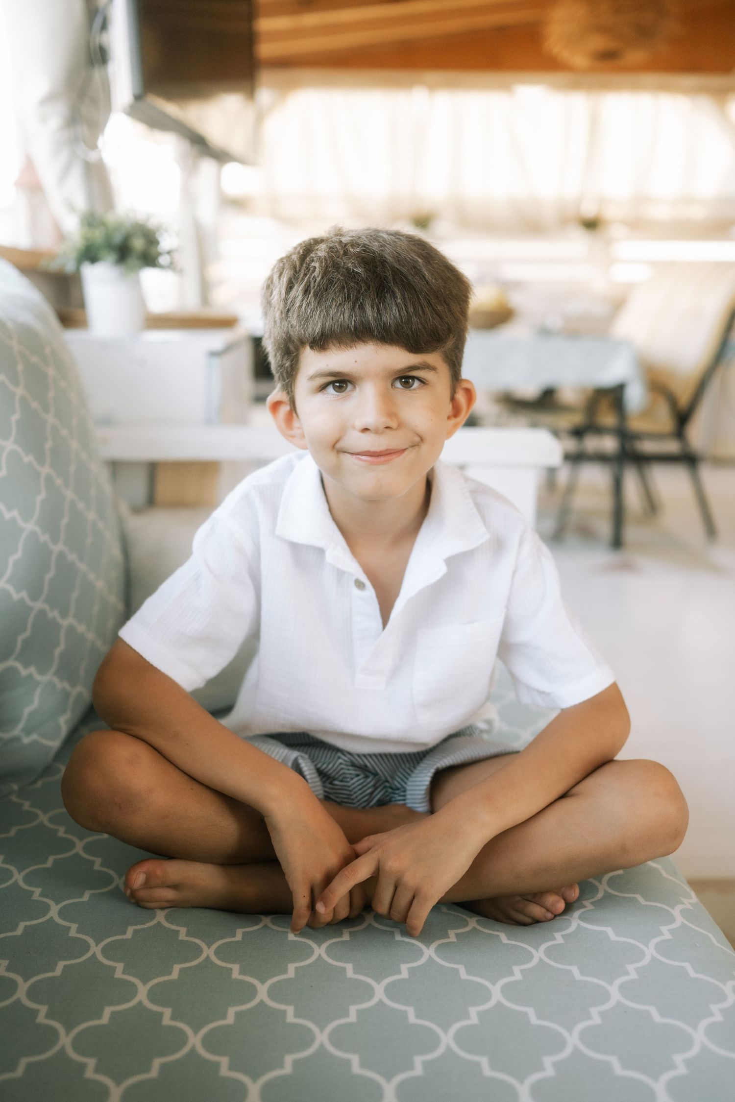 Child in white polo shirt sits cross-legged on gray patterned couch in candid lifestyle photo series.