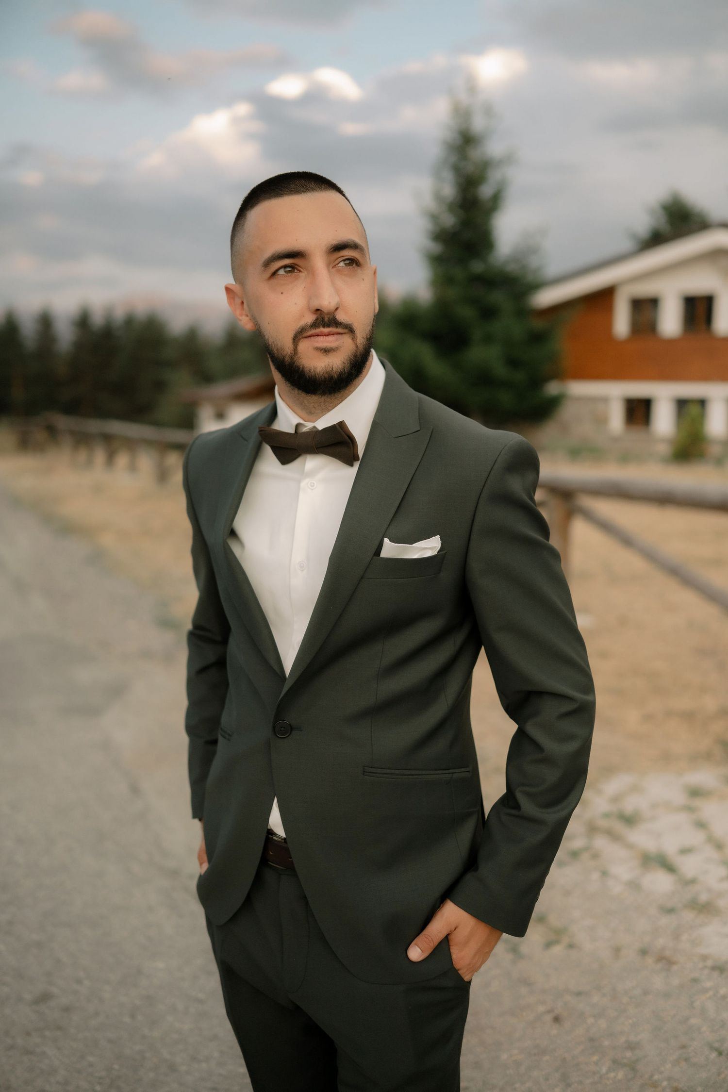 A person in a formal black tuxedo and bowtie poses outdoors against a cloudy sky and rustic background.