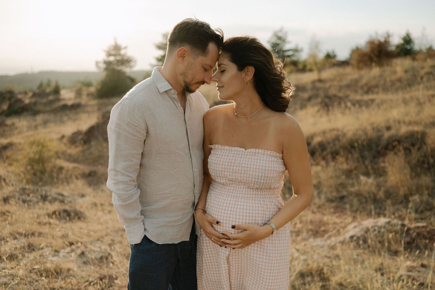 A romantic couple embraces in a golden field during sunset while wearing light colored outfits.