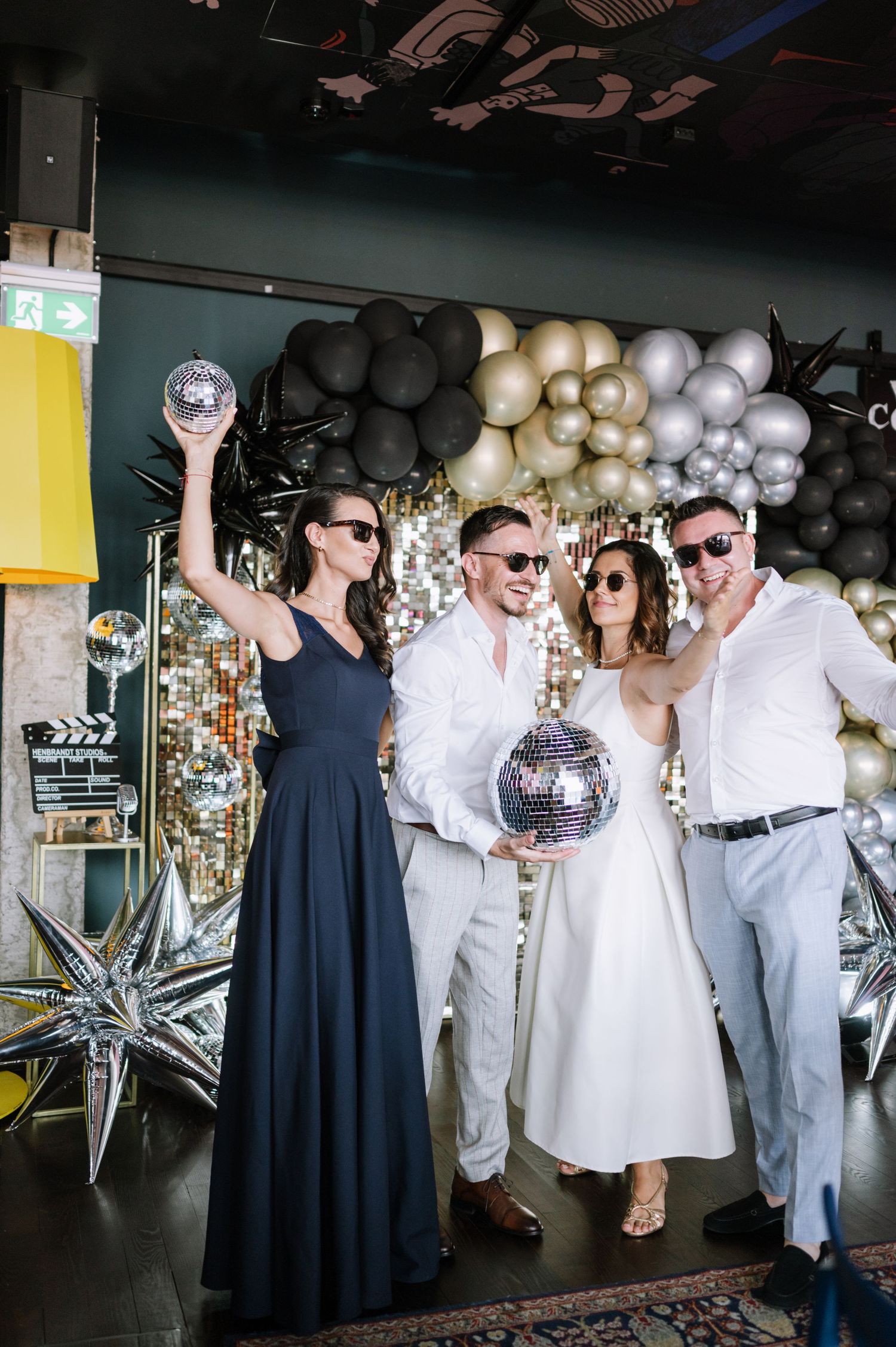 A group in navy dresses and white outfits pose playfully with disco balls and star decorations at a festive party venue.