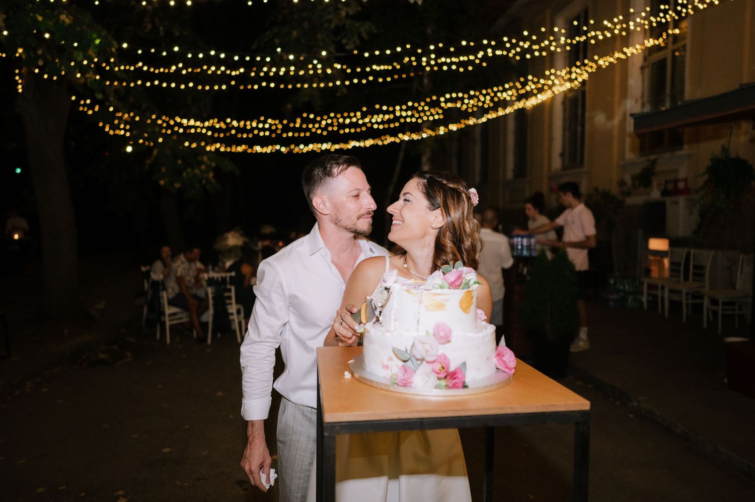 String lights illuminate an outdoor evening wedding reception with guests mingling at wooden tables.