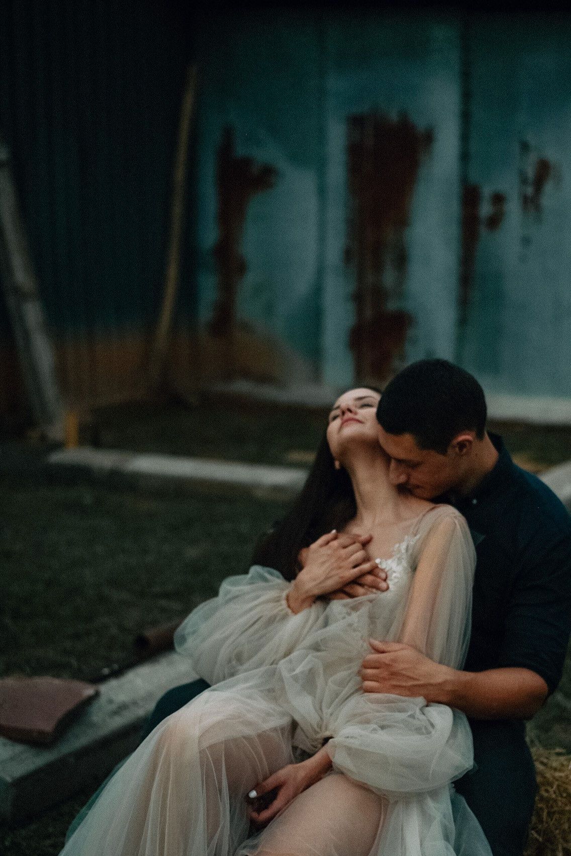 A romantic couple embraces and shares a tender kiss in a moody outdoor setting with a teal-colored wall background.