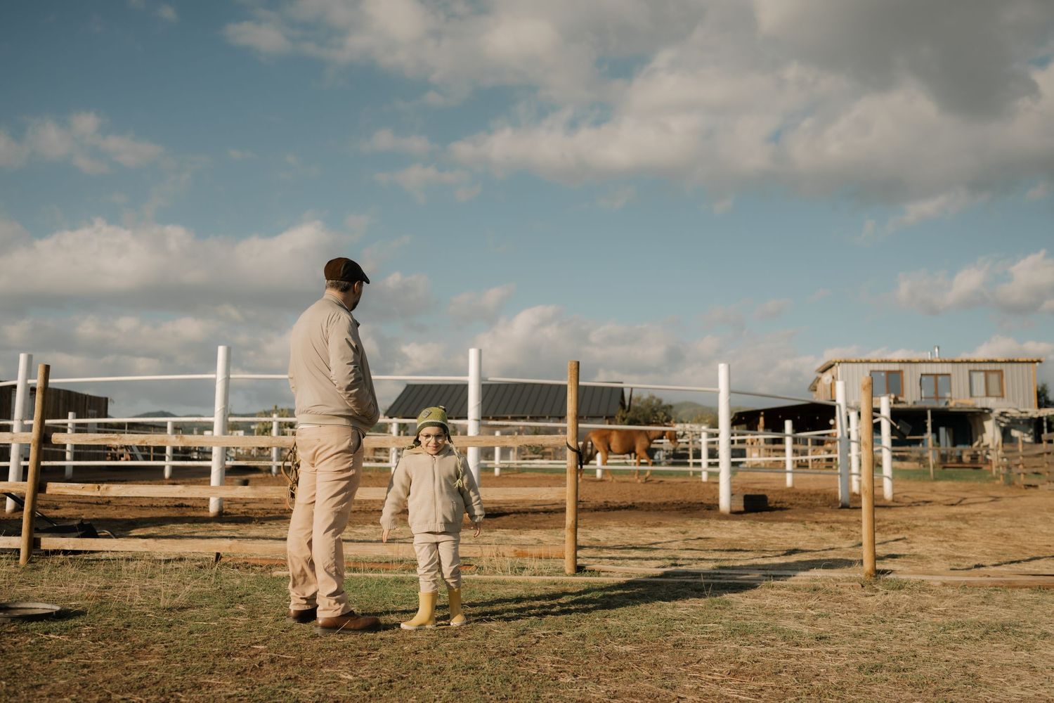 A panoramic view of a person in protective gear walking along white fenced paddocks on a ranch under cloudy skies.