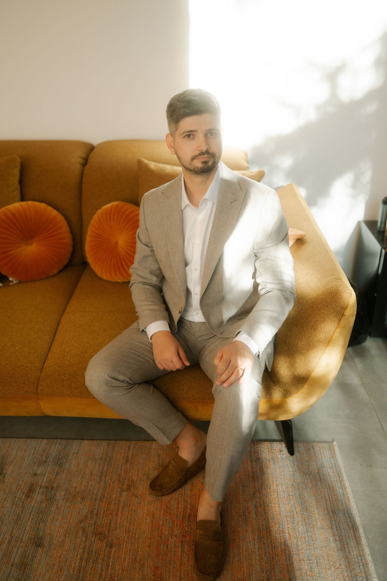 A person wearing a beige blazer and white shirt poses on an orange couch with round decorative pillows.