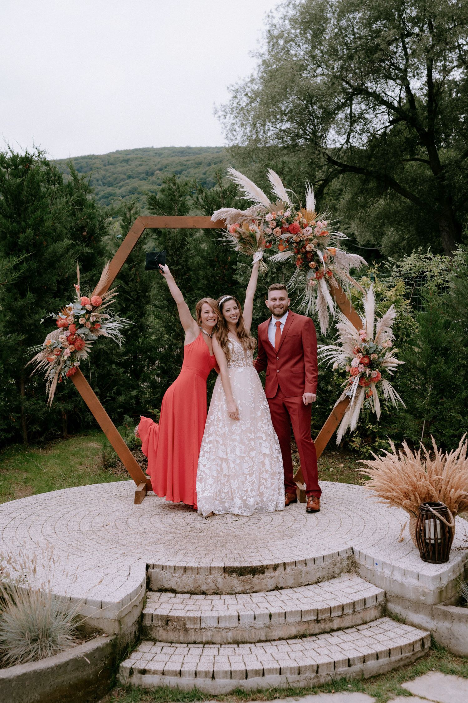 Wedding party celebrates on stone patio steps with hexagonal arch and dried floral arrangements in outdoor setting.