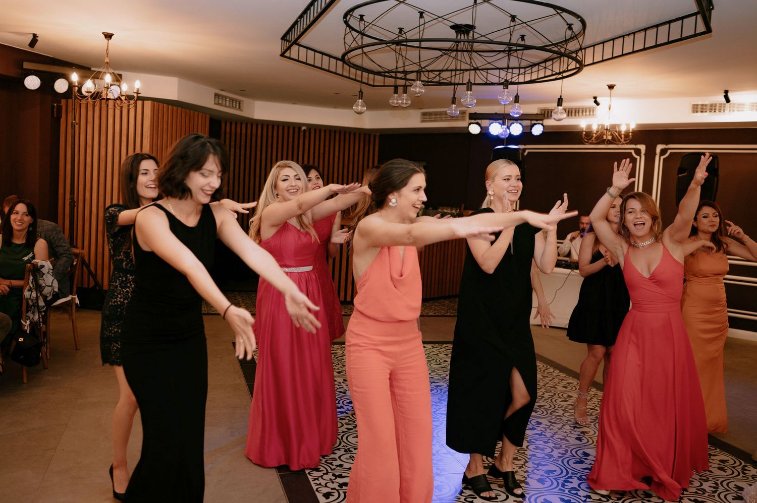 Wedding guests dance together in a reception hall with circular lighting fixtures and wood paneled walls.