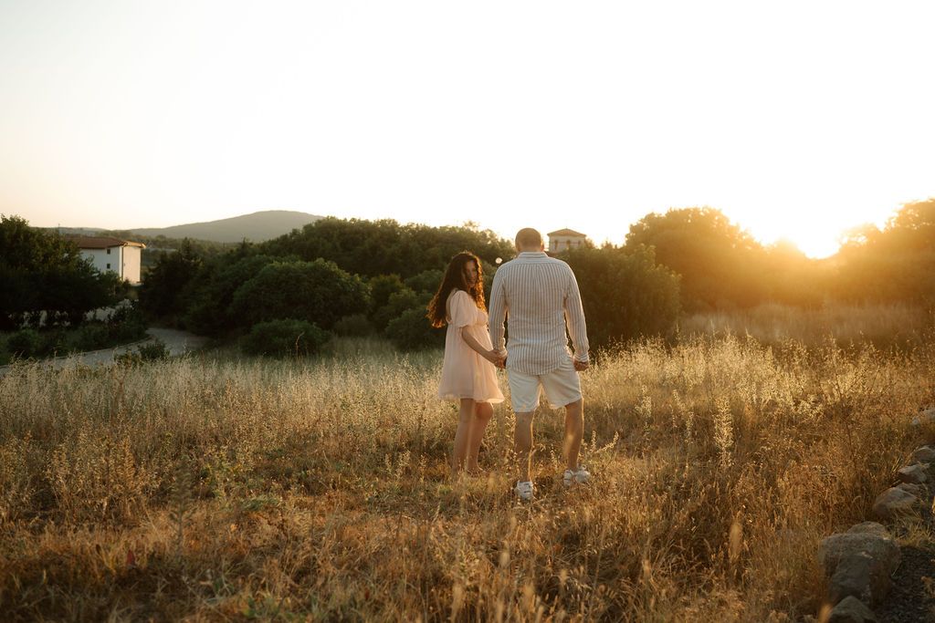 A couple walks through a golden field at sunset with mountains in the background.
