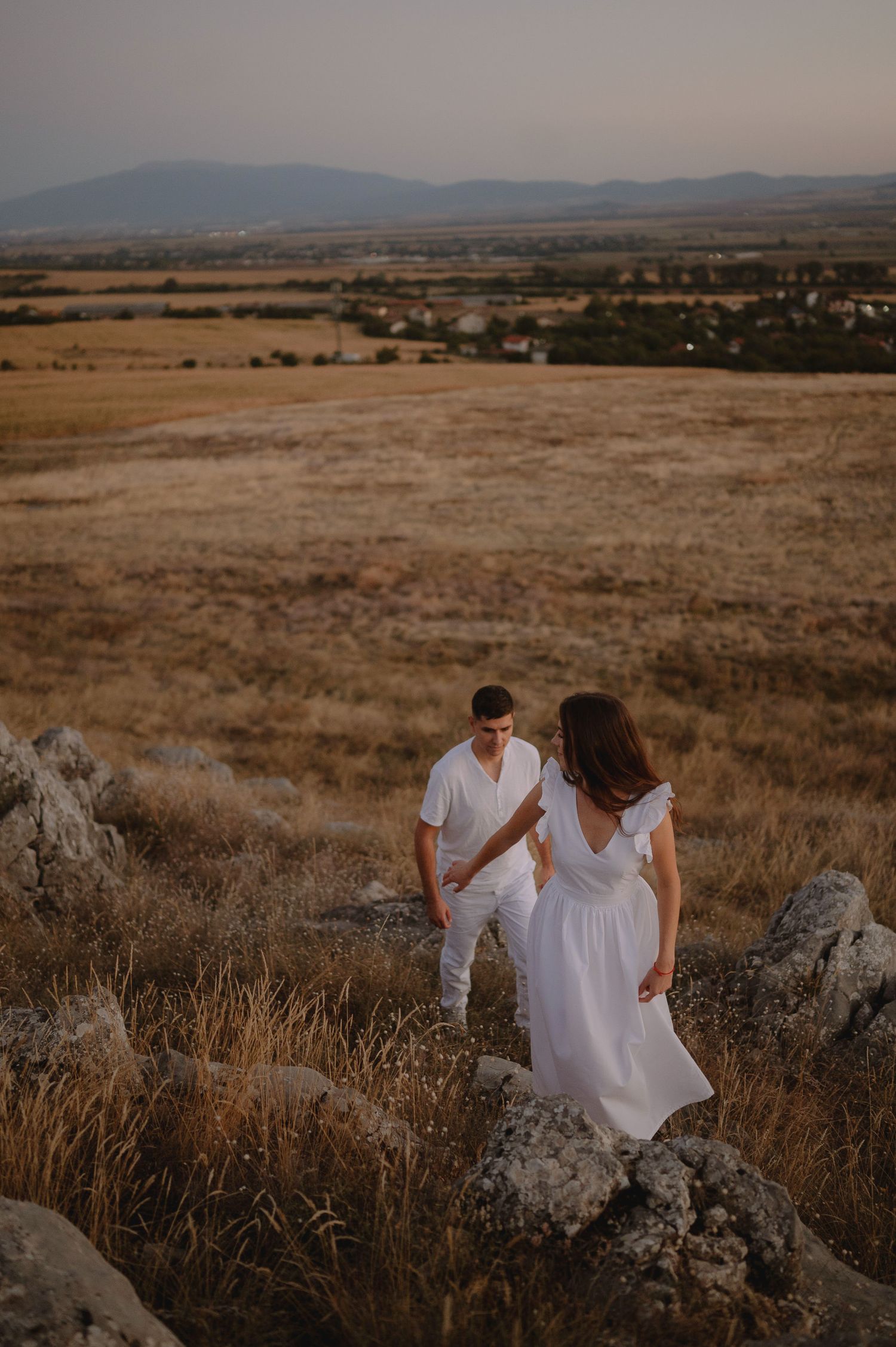 Couple in white outfits walking through golden grasslands with mountains in the background at sunset.