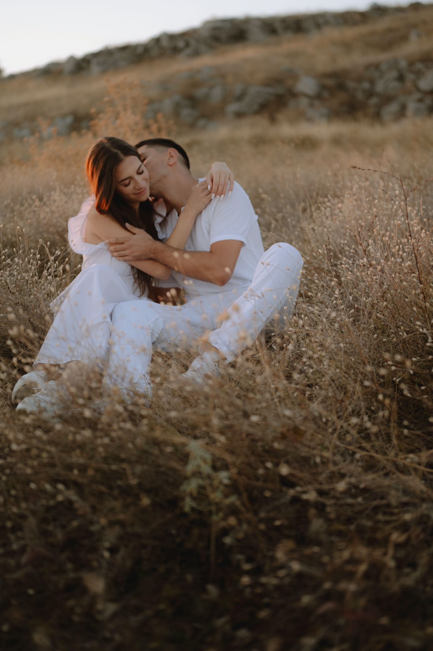 Couple in white outfits share intimate moments while lying in a golden field during sunset.