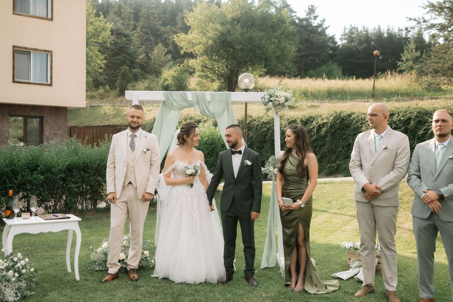 Wedding party members in light colored suits and dresses pose together on a grassy lawn during an outdoor celebration.