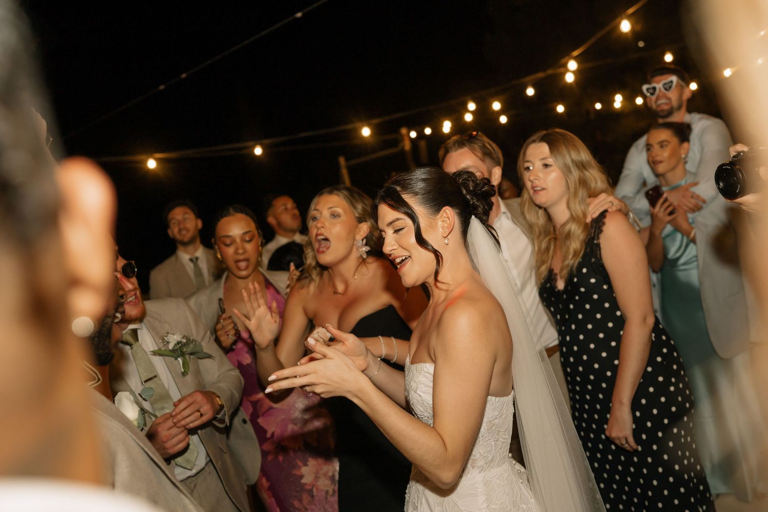 Bride and guests laughing and celebrating together at an outdoor wedding reception under string lights at night.