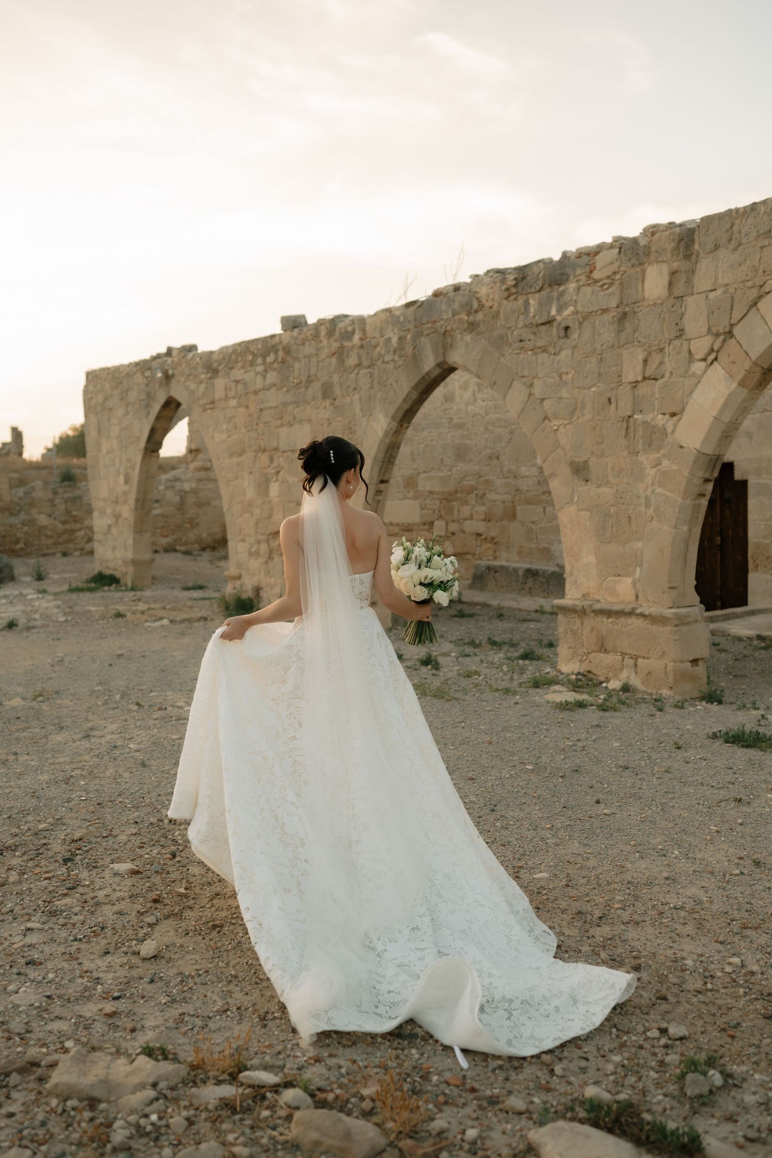 Bride in white lace gown holding bouquet stands before ancient stone arches ruins at sunset.