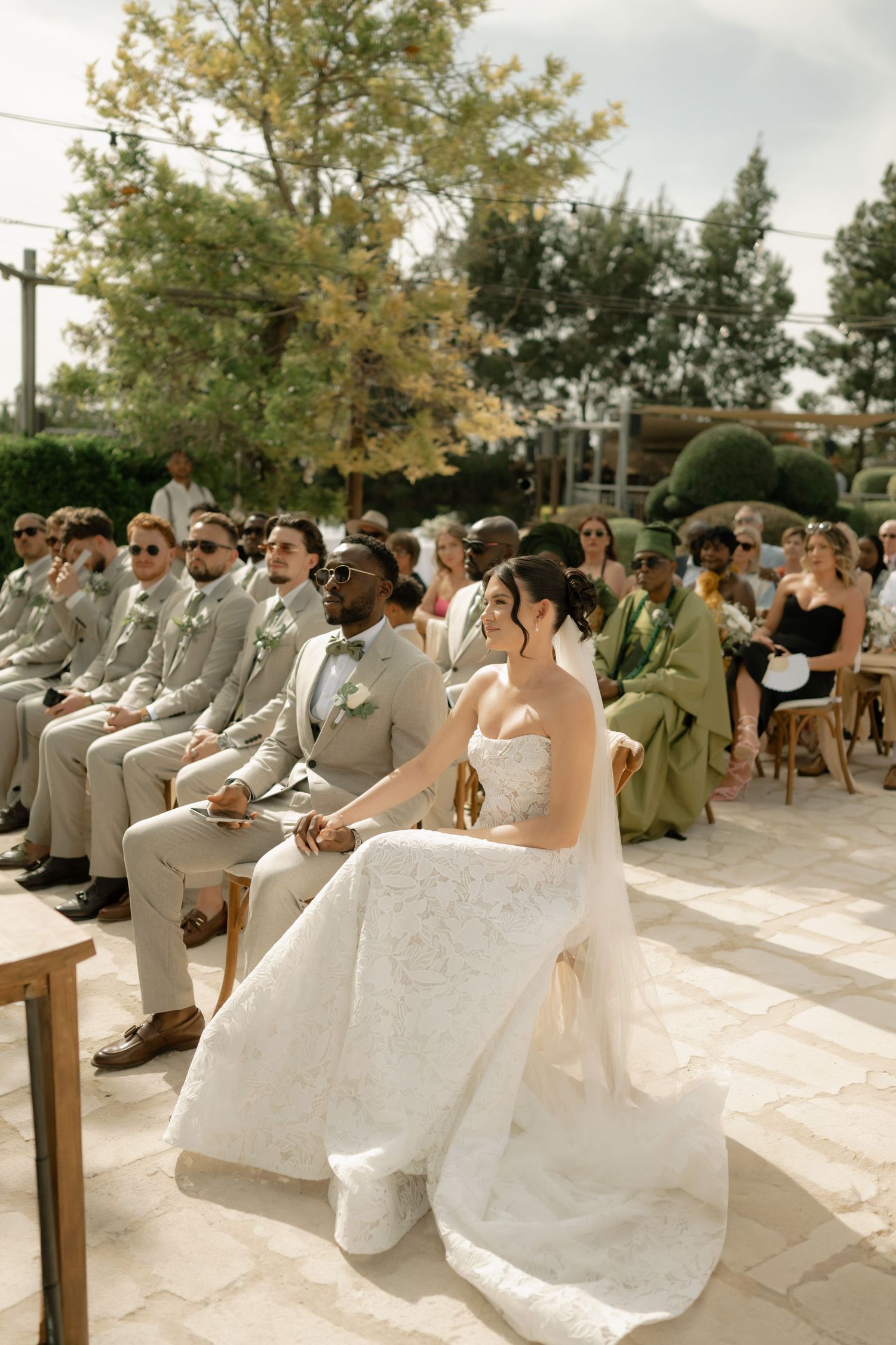 Bride in white gown and groom in gray suit seated at outdoor wedding ceremony with guests in background.