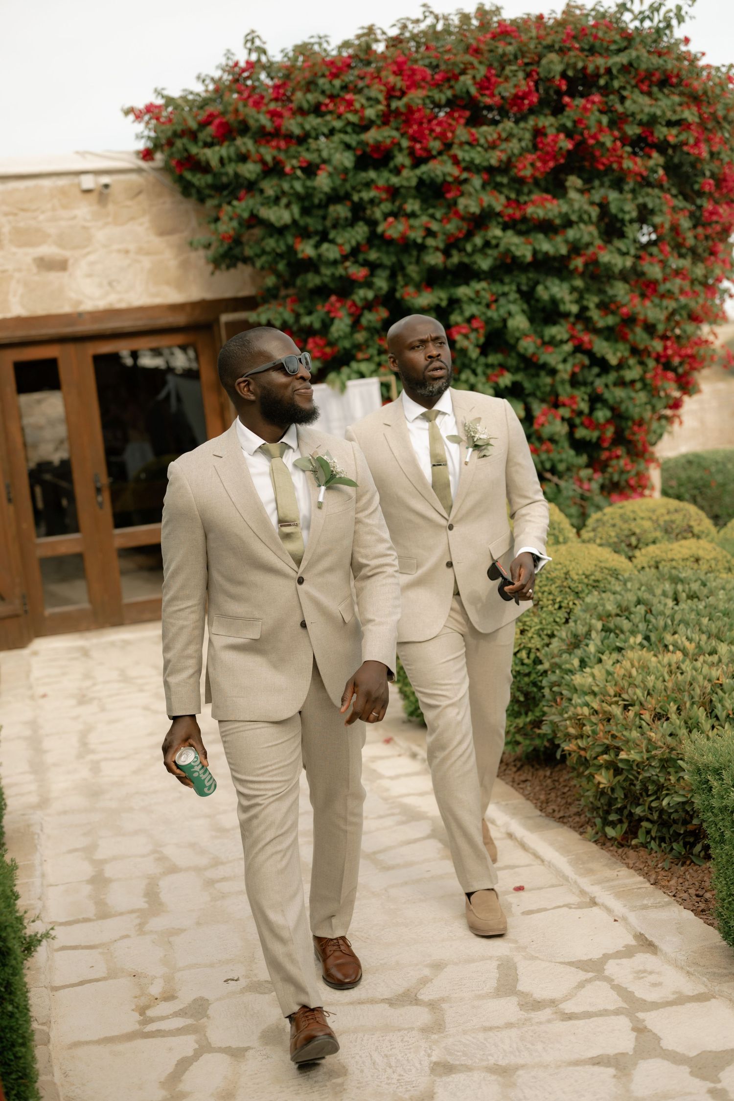 Two groomsmen in matching tan suits with boutonnieres walk along a stone path beside a flowering red bougainvillea.