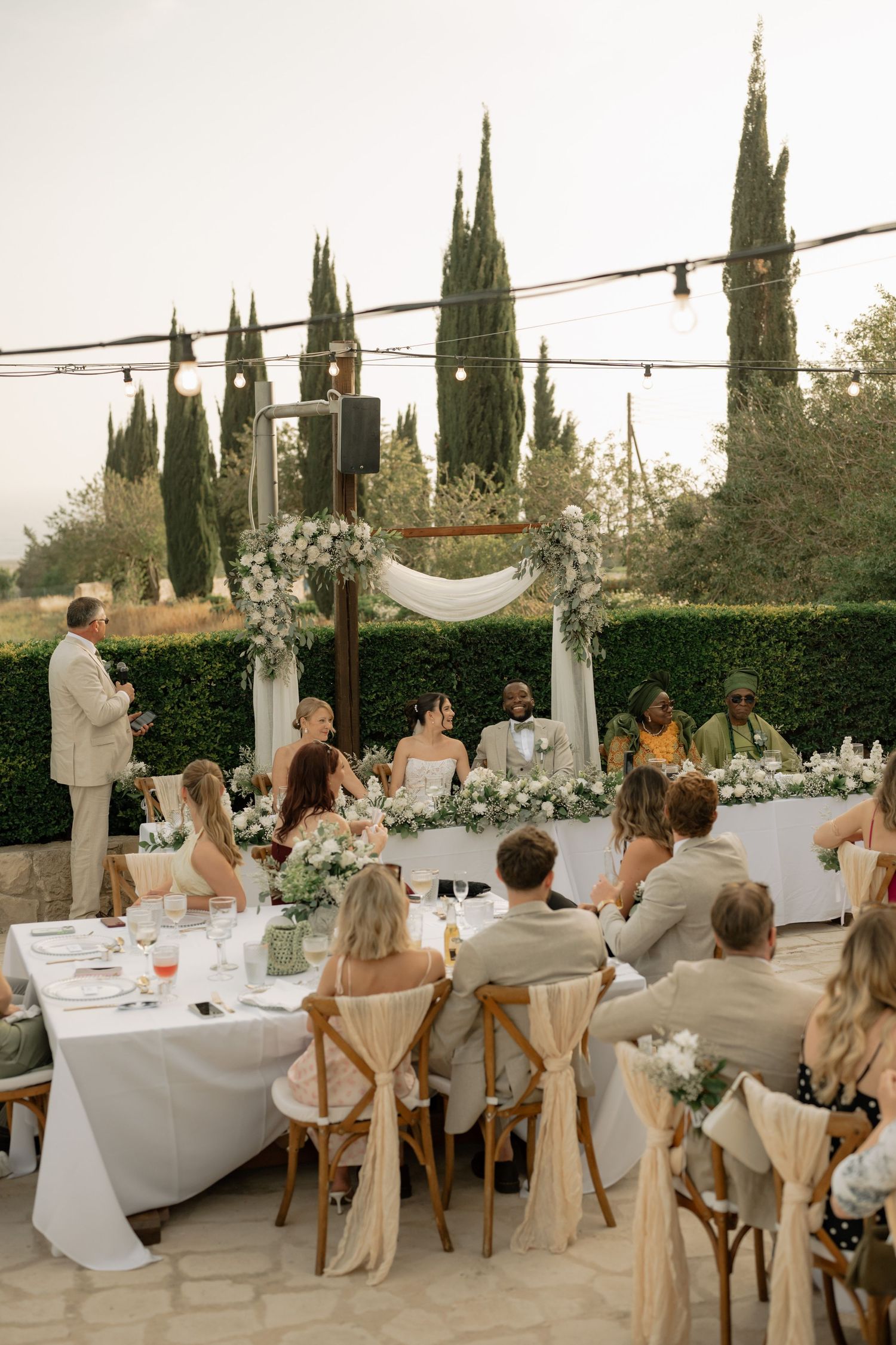 Elegant outdoor wedding reception with floral arch, string lights, cypress trees, and guests seated at white-draped tables.