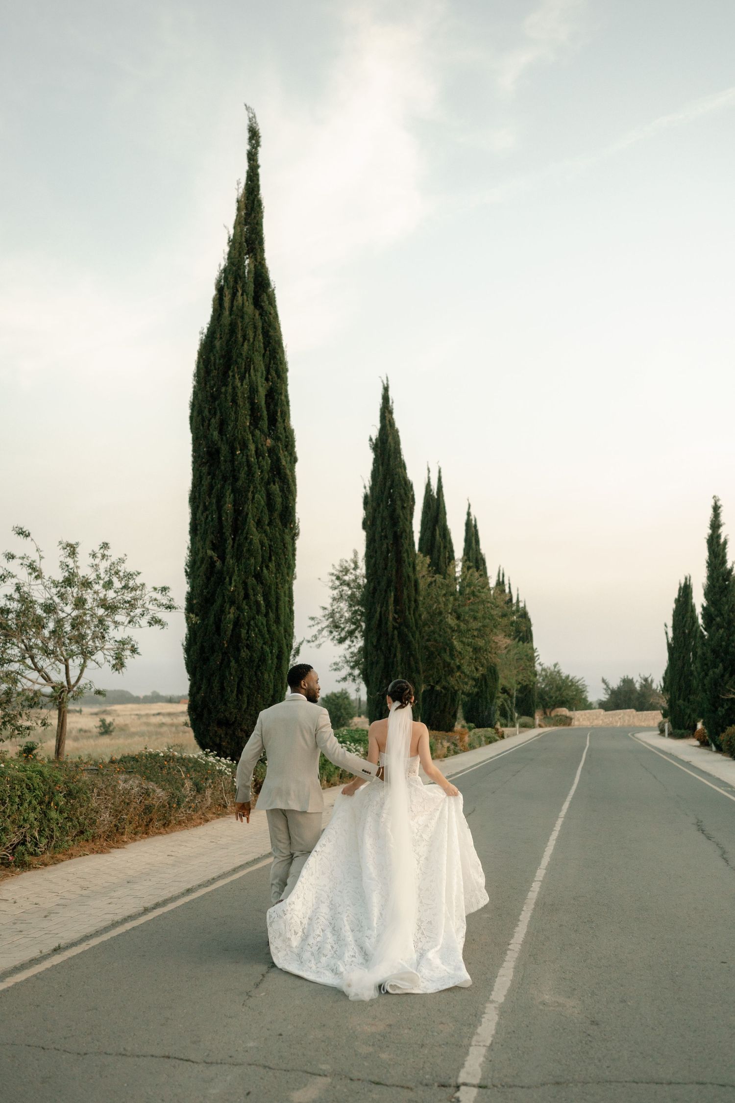 Bride and groom walking hand in hand down a tree-lined road on their wedding day.