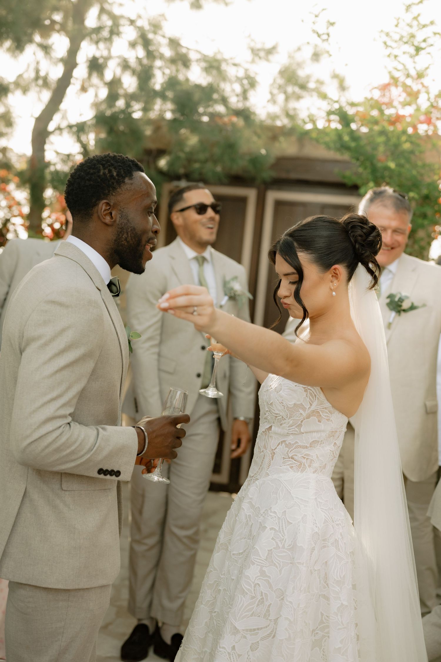 Bride and groom clinking champagne glasses at outdoor wedding reception with groomsmen in beige suits in background.