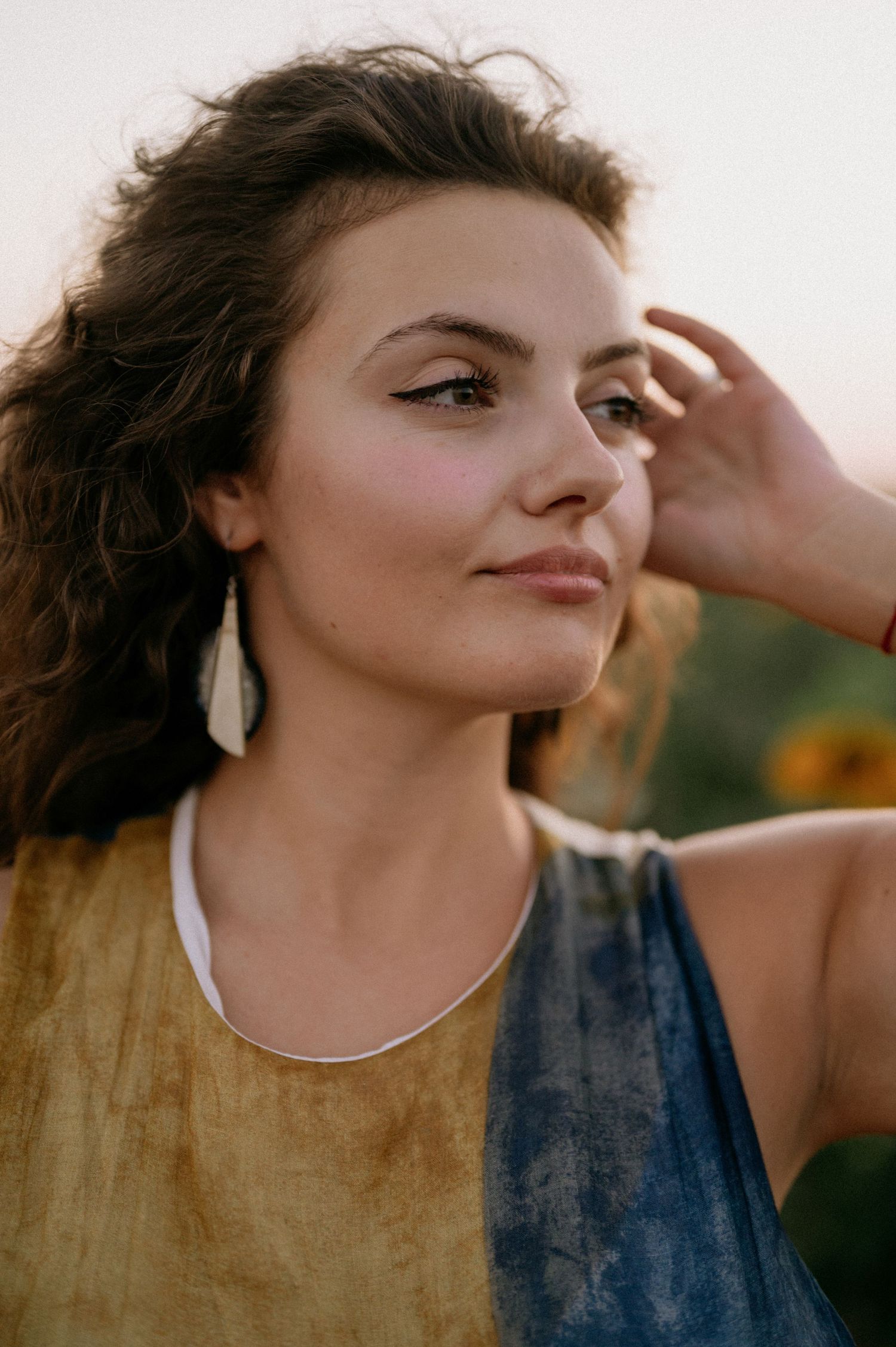 Young woman with curly hair wearing a two-tone dress and white earrings, posing outdoors in golden hour light.