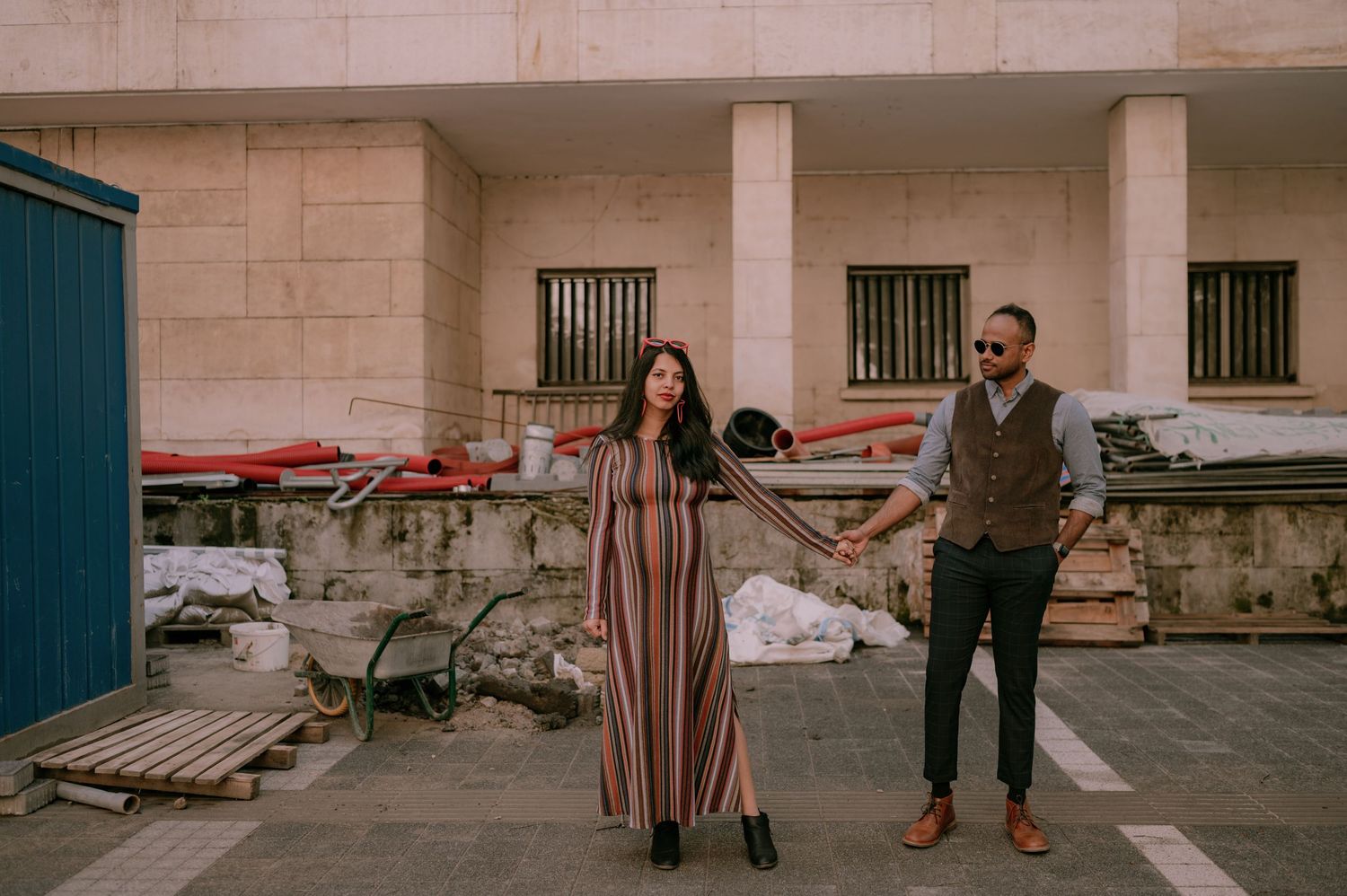 Couple holding hands in striped dress and vest outfit posing outdoors near stone building with construction materials.
