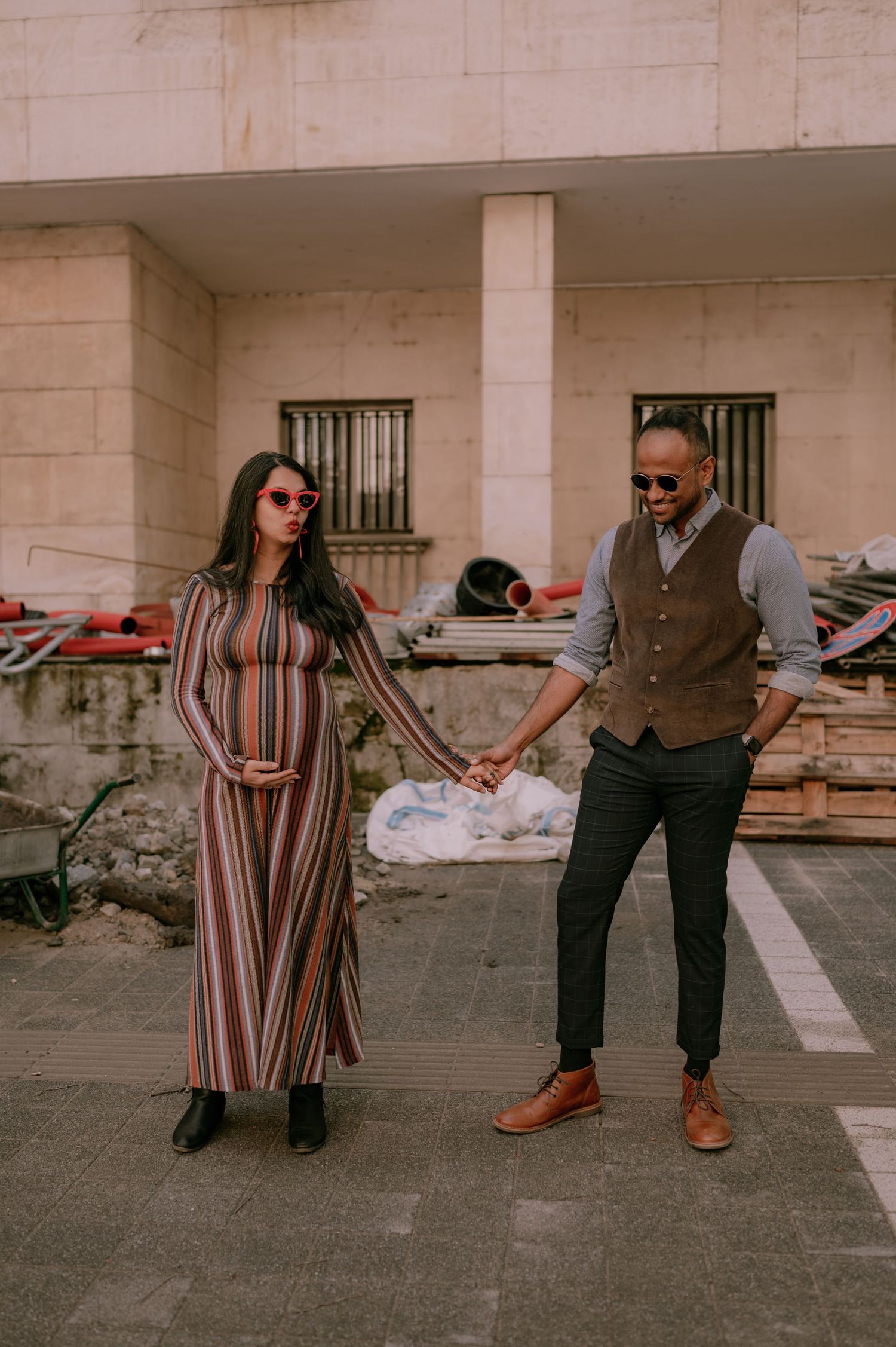 Pregnant woman in striped maxi dress holds hands with stylish man in vest and sunglasses outdoors.