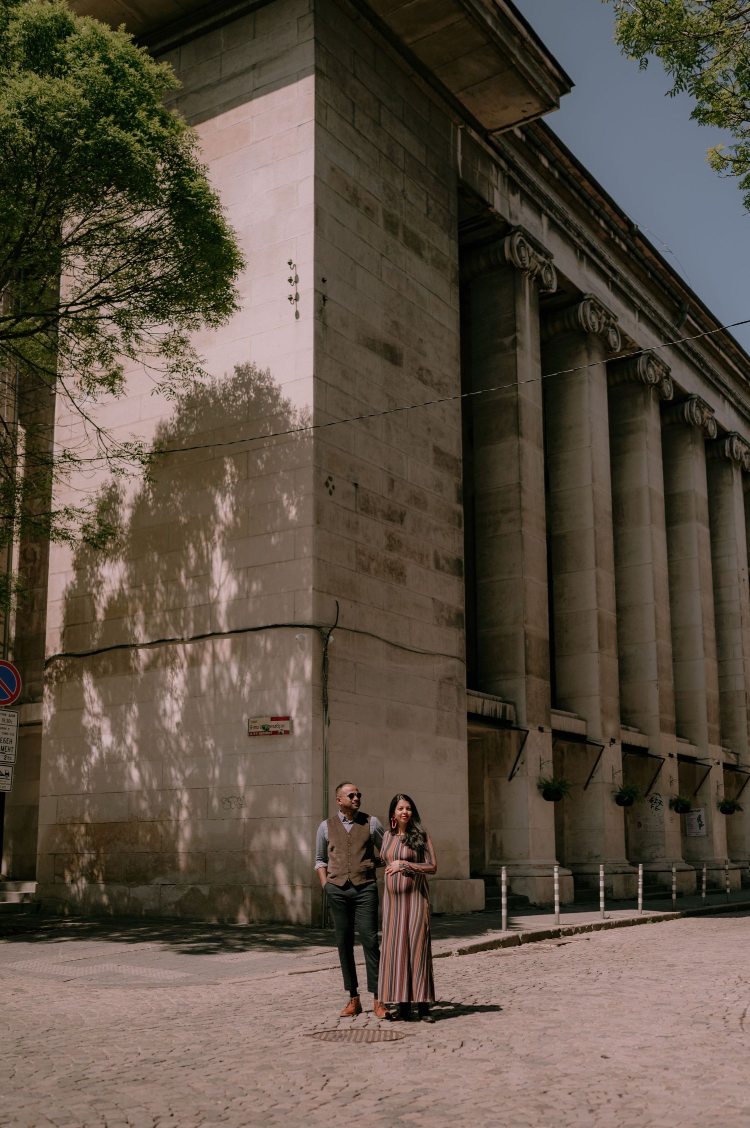 Couple standing near a grand neoclassical building with tall columns on a sunny cobblestone street.