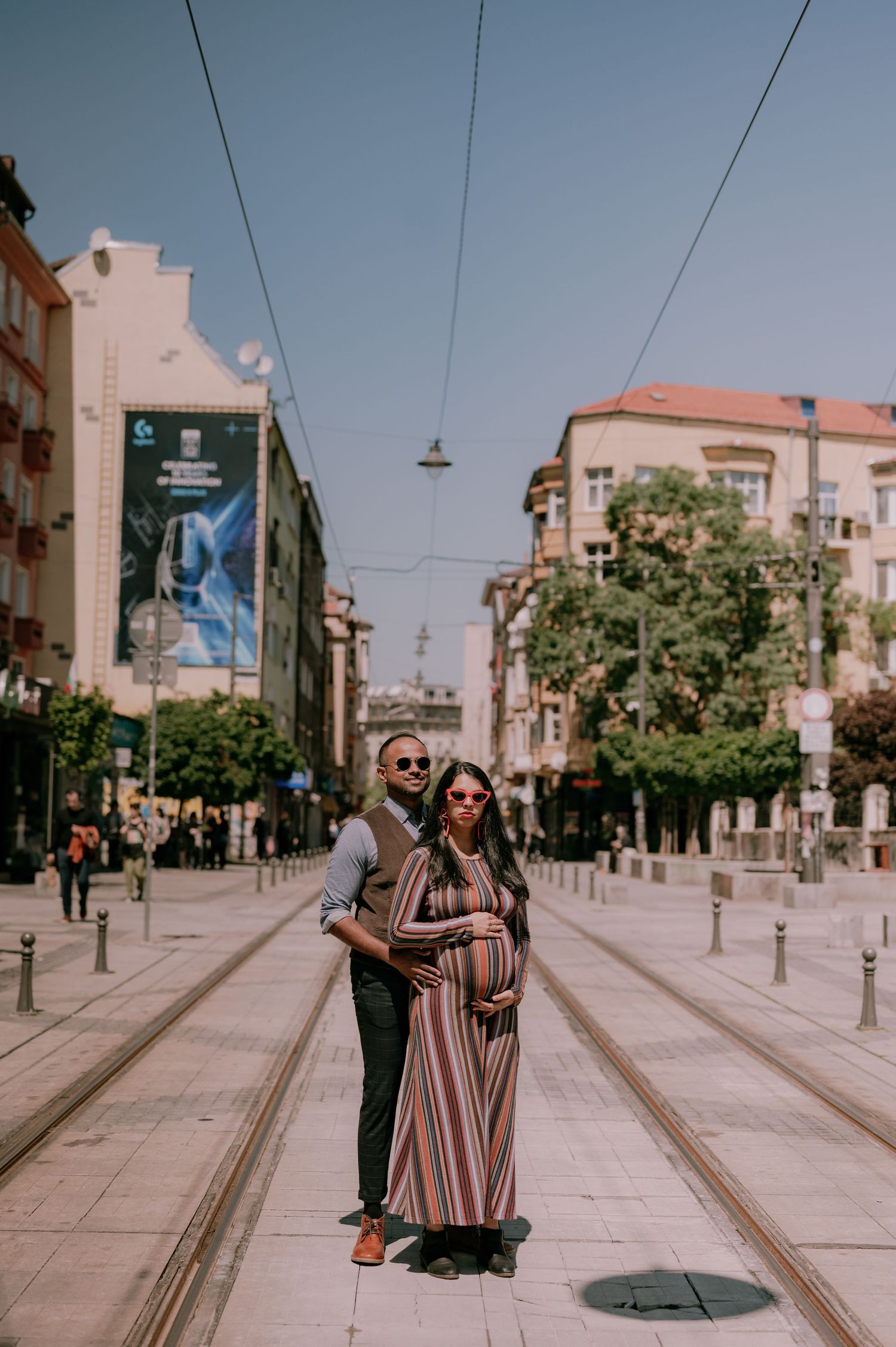 A pregnant couple poses on a tram street in an urban European city, the woman in a striped maxi dress cradling her baby bump.