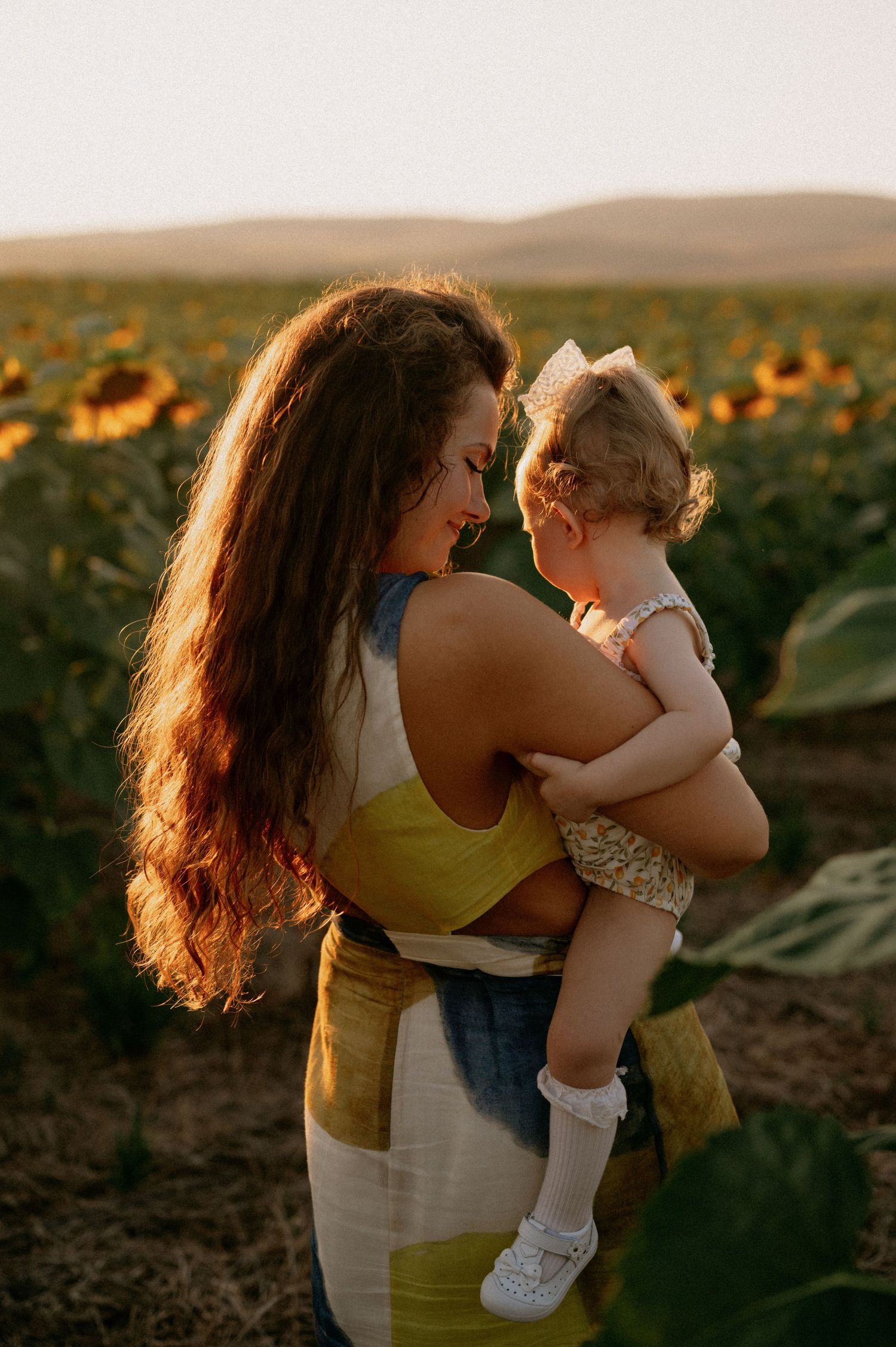A mother holds her toddler in a sunflower field at golden hour sunset.