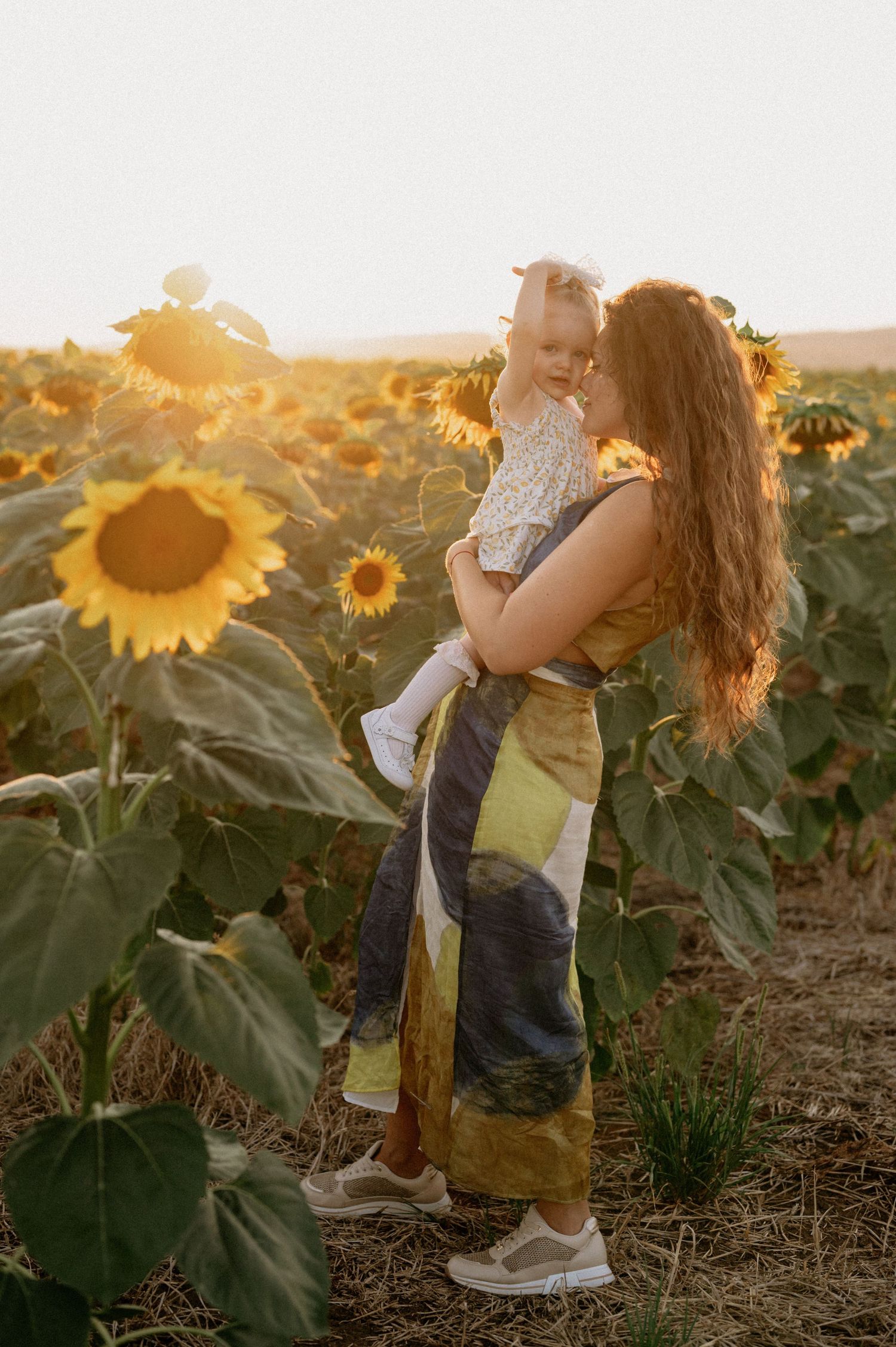 A mother holds her toddler in a golden sunflower field at sunset, both smiling in a warm, glowing outdoor portrait.