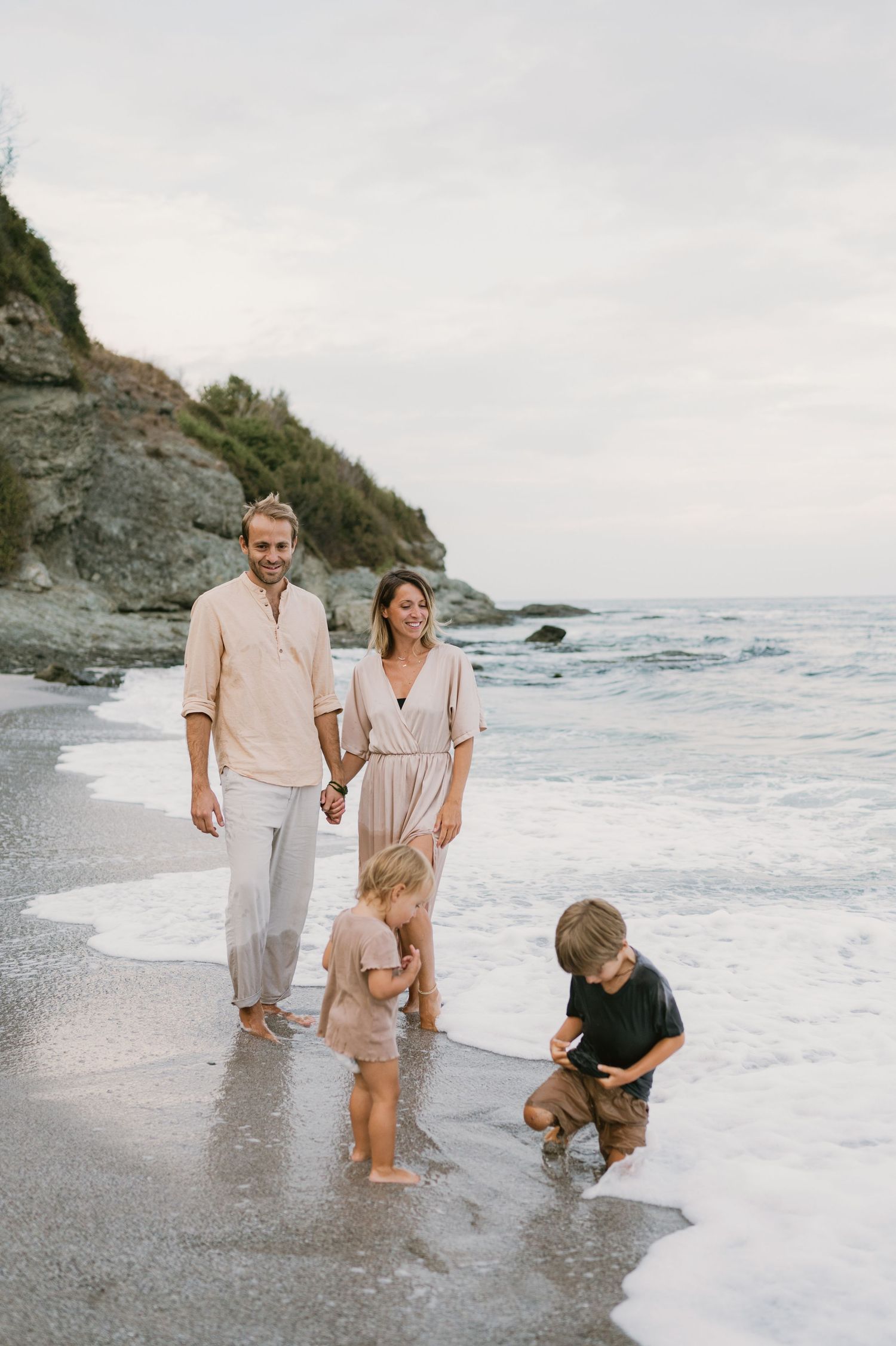 Family of four enjoying a beach walk, parents holding hands while children play in ocean waves near a rocky cliff.