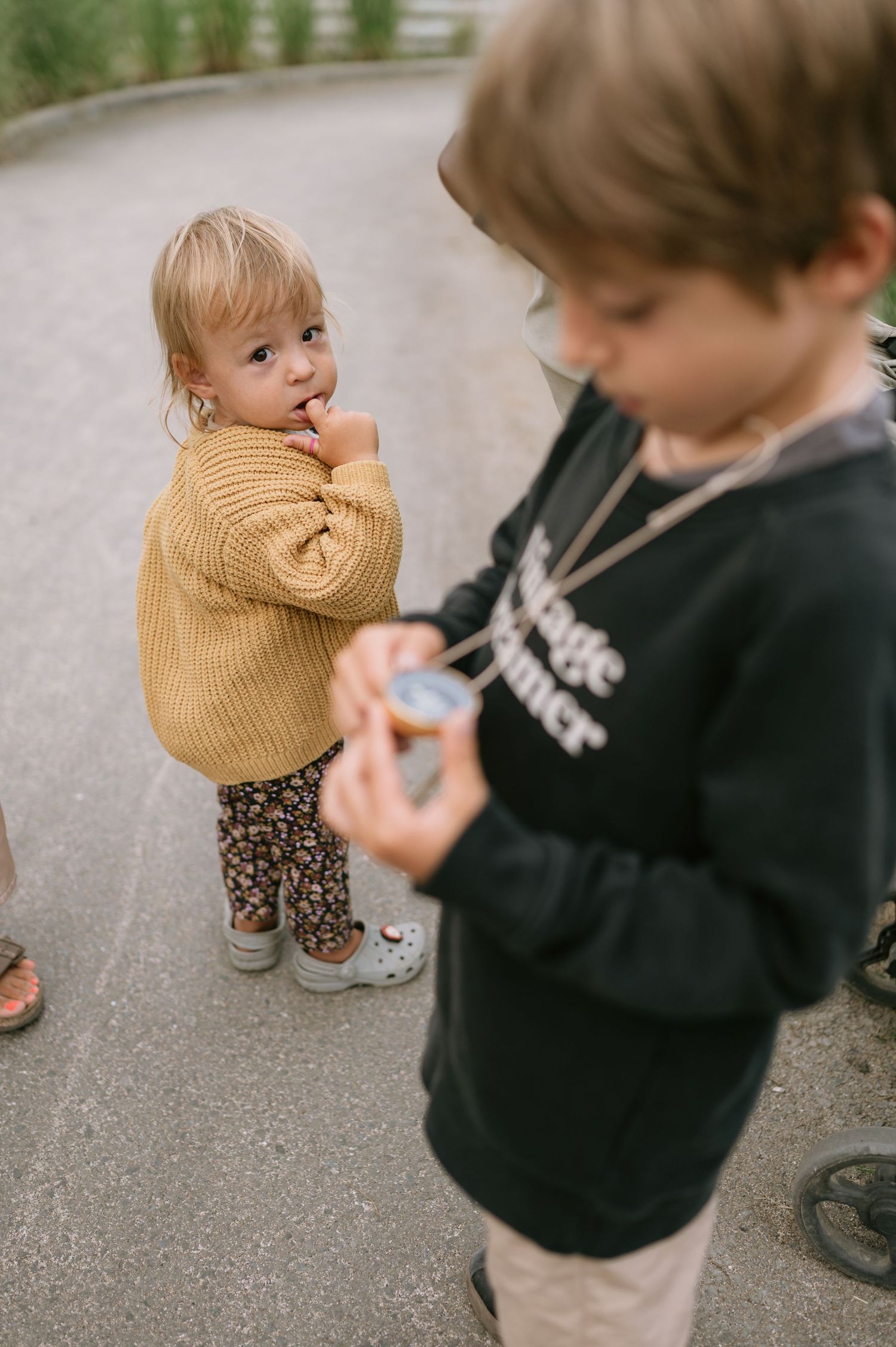 Two young children outdoors, toddler in yellow sweater and floral pants, older child in black sweatshirt holding small object.