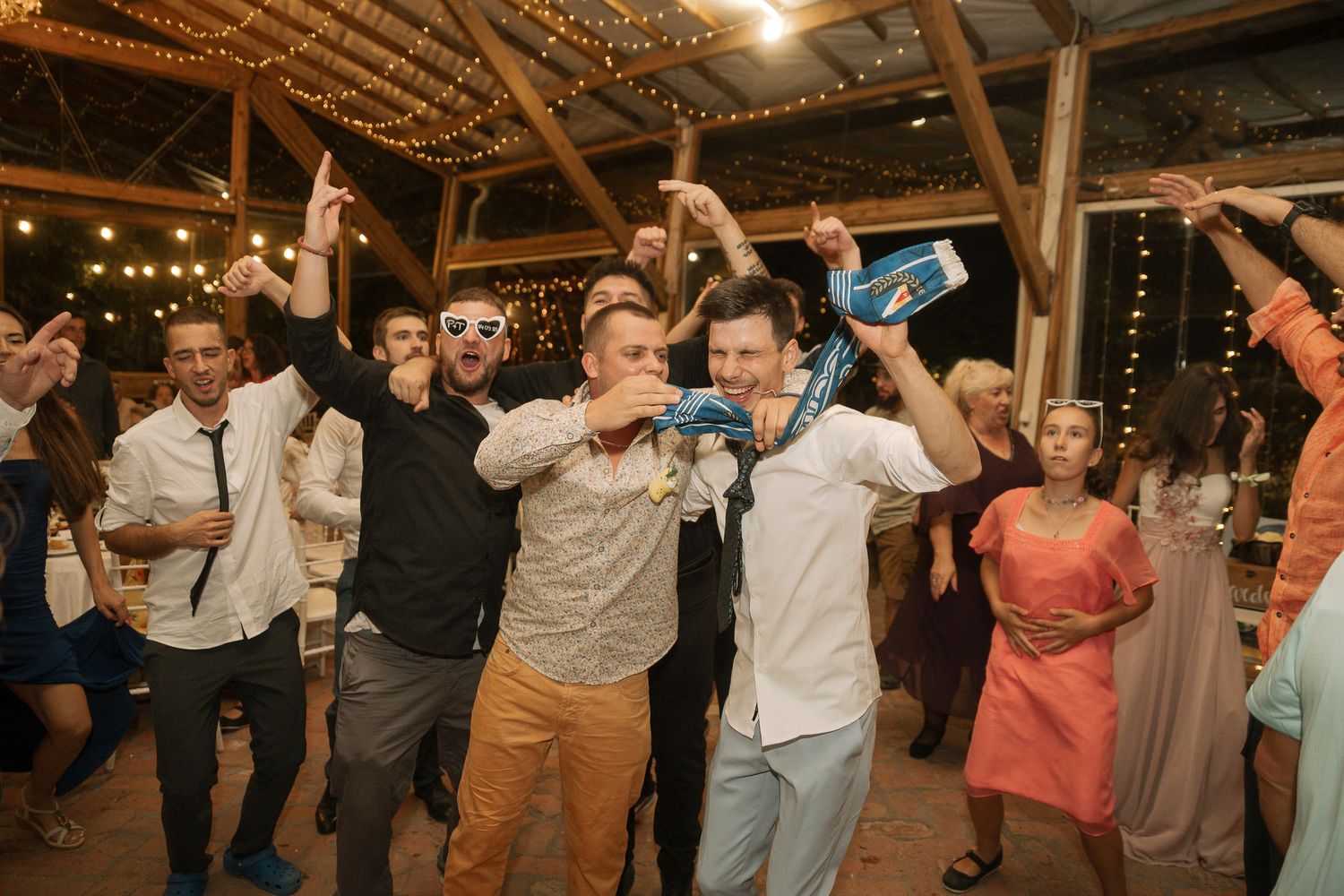 A group of wedding guests dancing and celebrating on a wooden floor under warm indoor lighting at a reception venue.