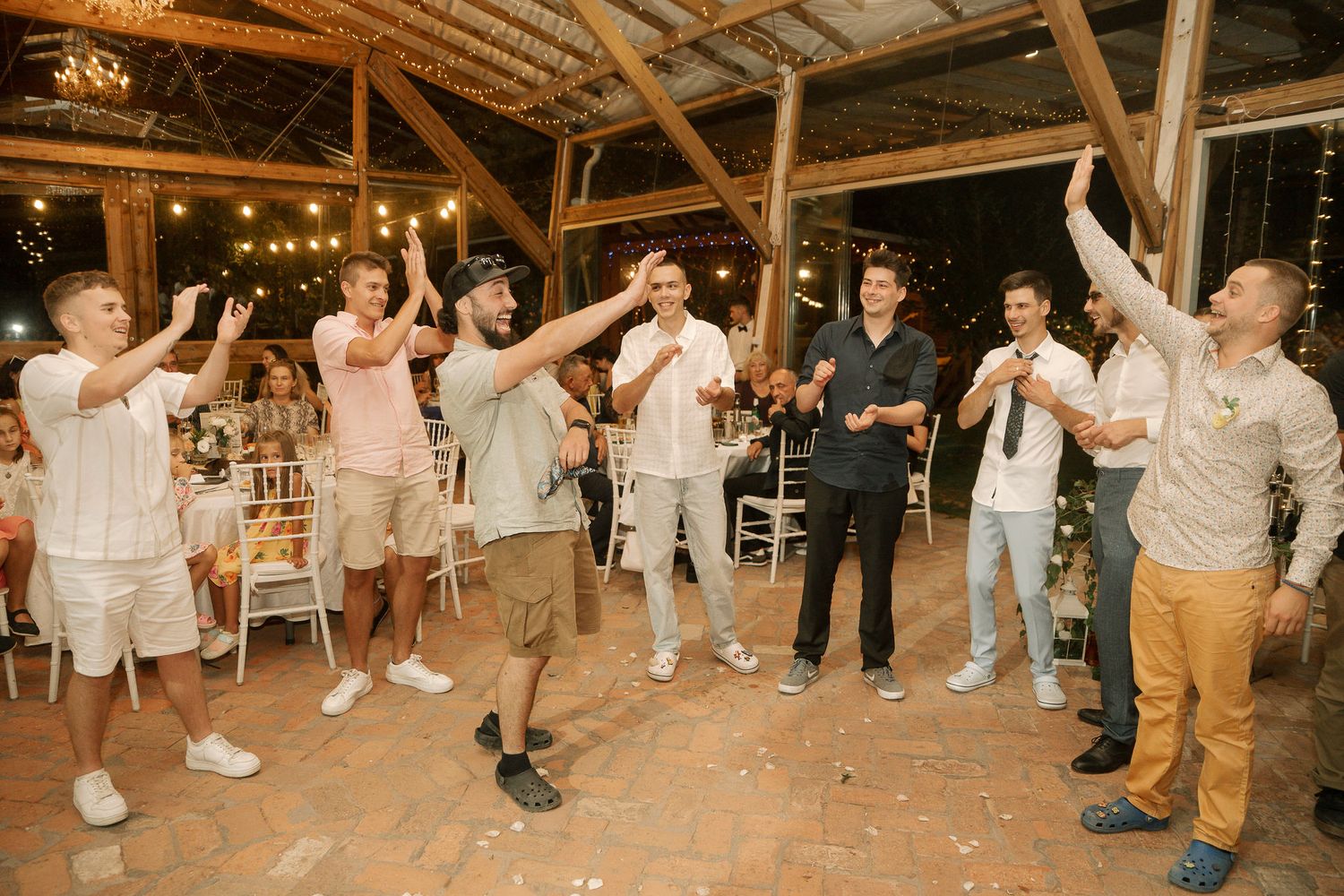 Wedding guests dance and celebrate in a rustic barn venue with wooden beams overhead during a lively reception party.