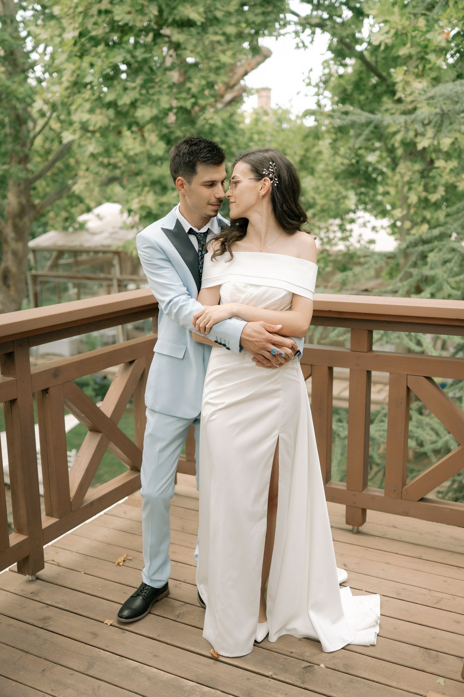A romantic wedding couple embraces on a wooden deck surrounded by lush green trees on their special day.