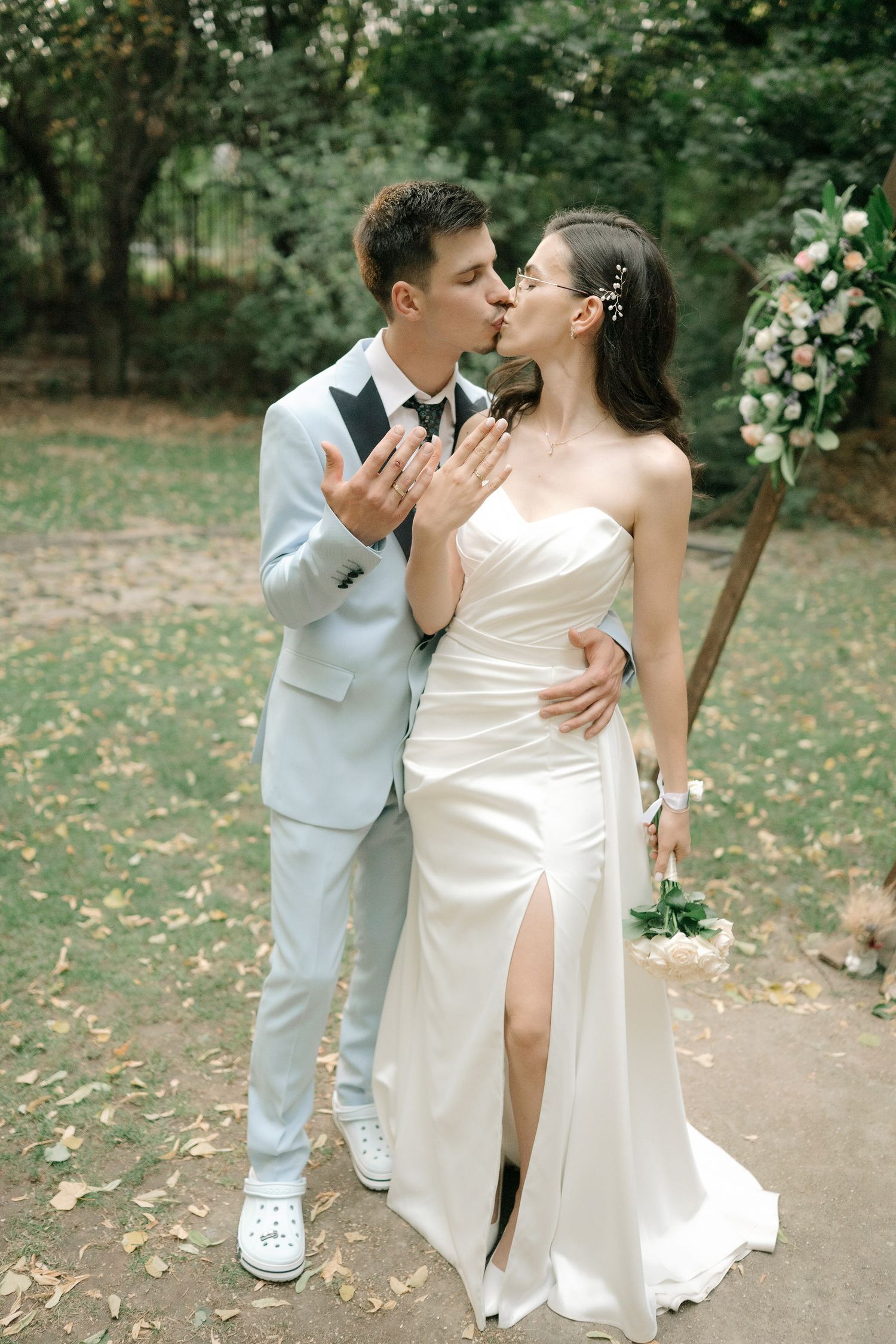 A couple in wedding attire share an intimate moment in a garden setting, with the bride in a slit dress and groom in light suit.