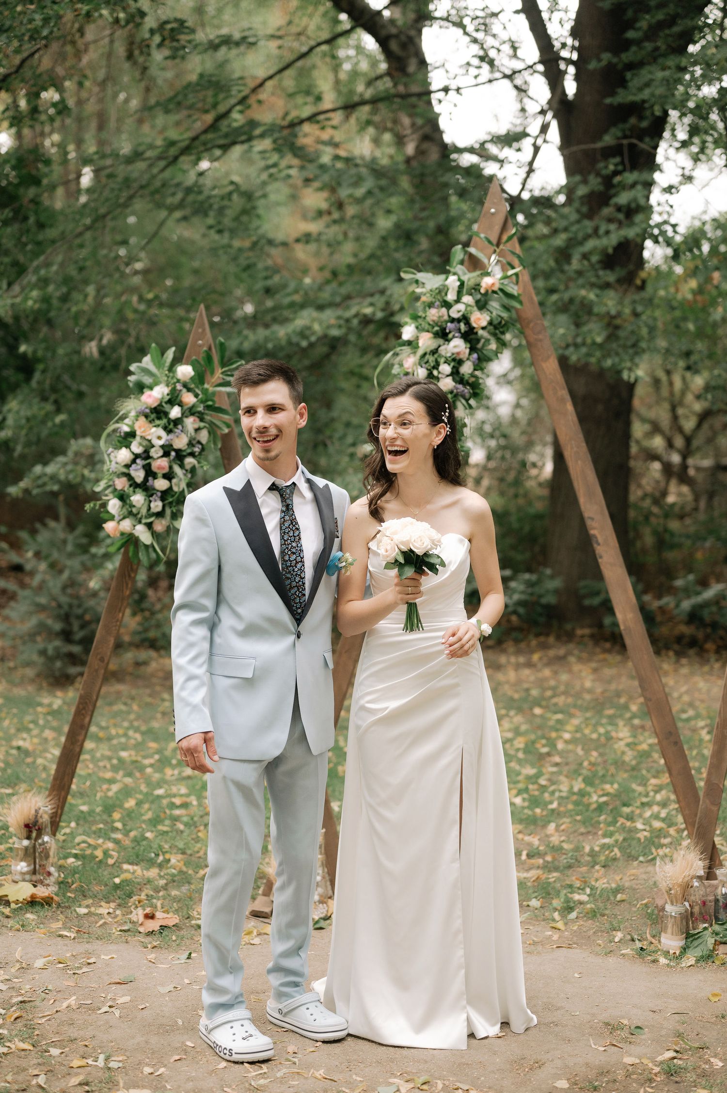 A joyful outdoor wedding ceremony with wooden arches decorated in white floral arrangements in a natural forest setting.