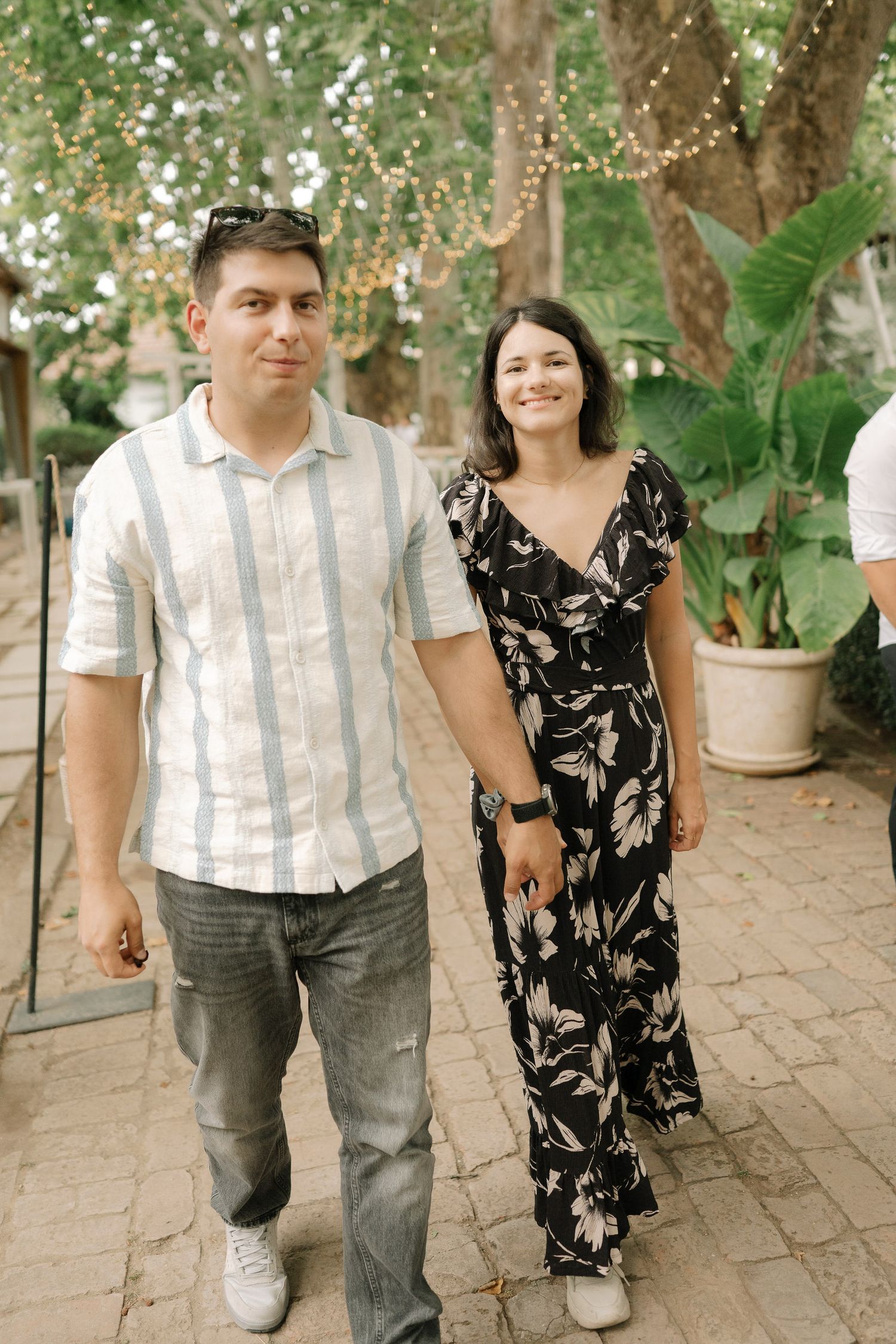 A couple walks hand in hand on a brick patio surrounded by lush greenery and potted plants.