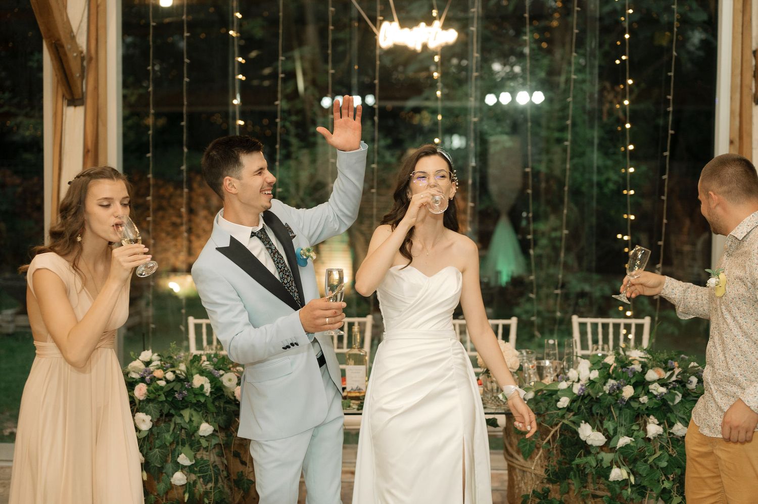 Wedding party celebrates at an elegant indoor reception with string lights and floral decorations.
